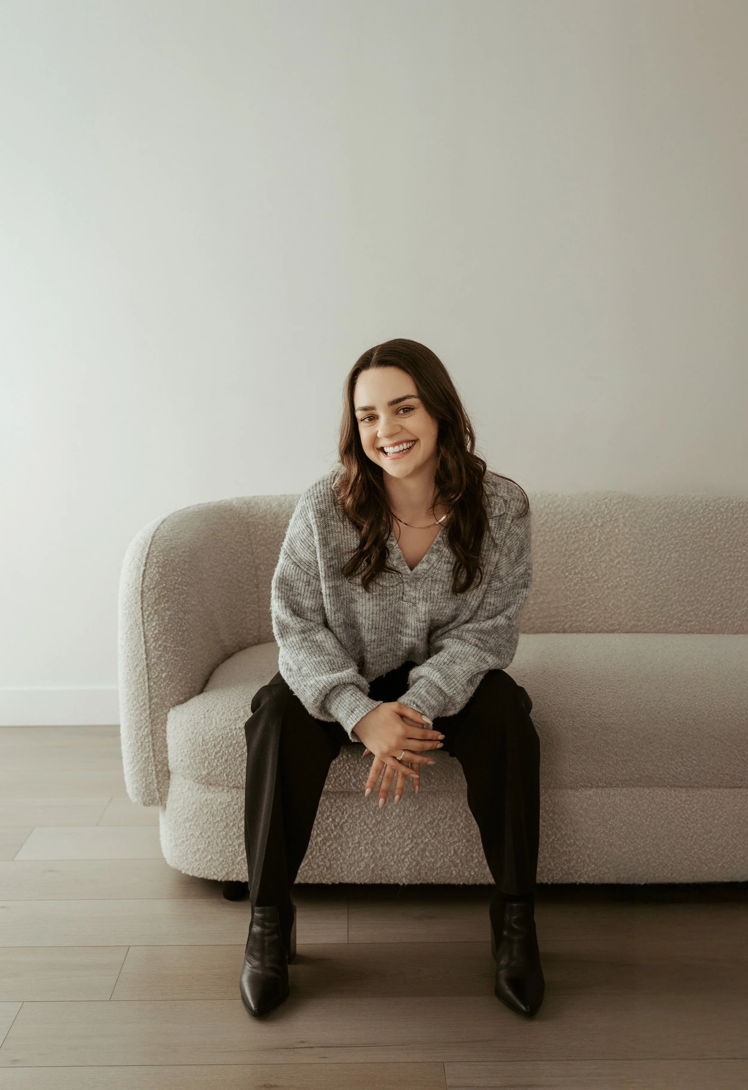 Short brown haired online counsellor leaning forward sitting on a white sofa, smiling at the camera. 