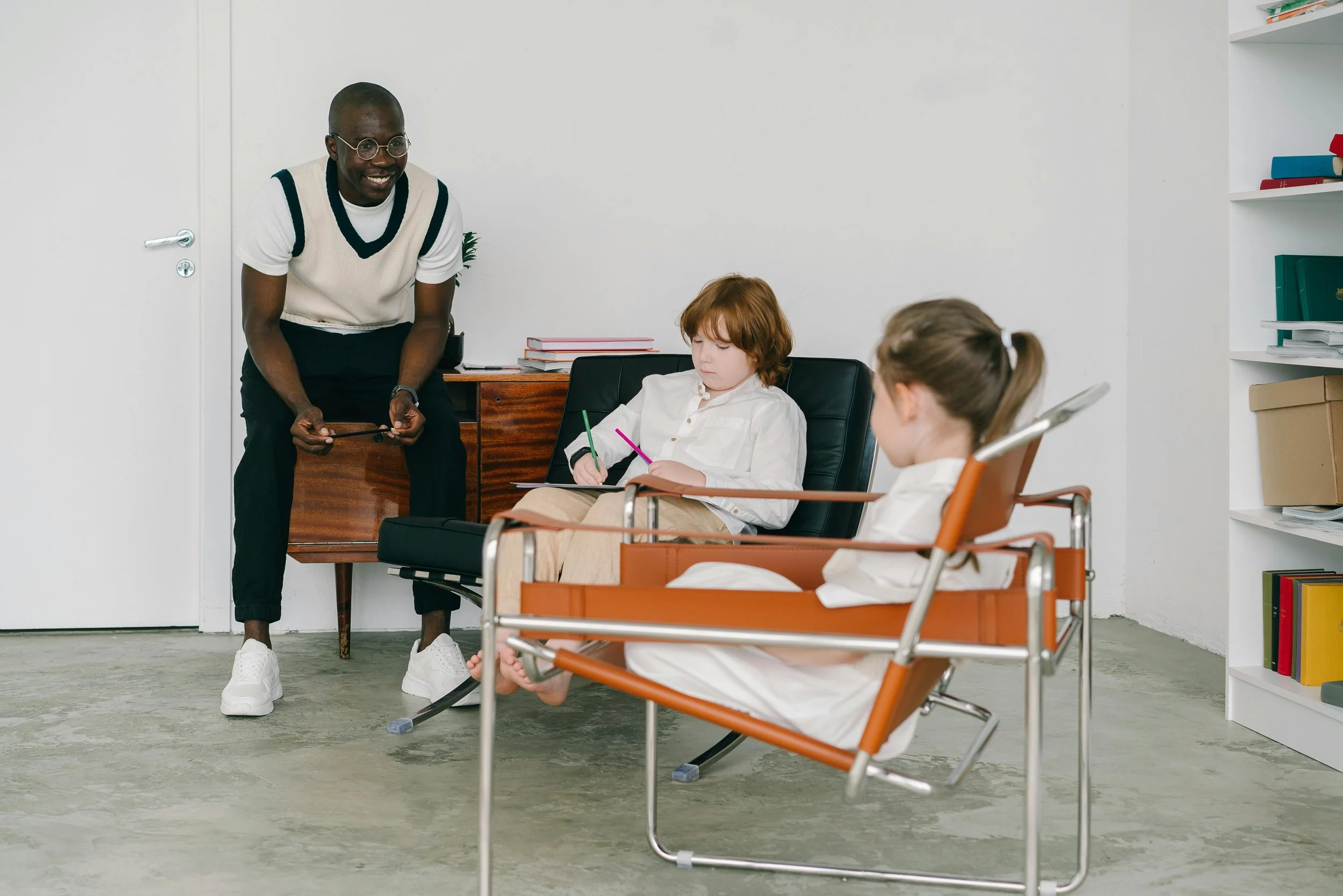 A man is standing and smiling, engaging with two children sitting in chairs, one in a wheelchair, in a room with white walls and a bookshelf.