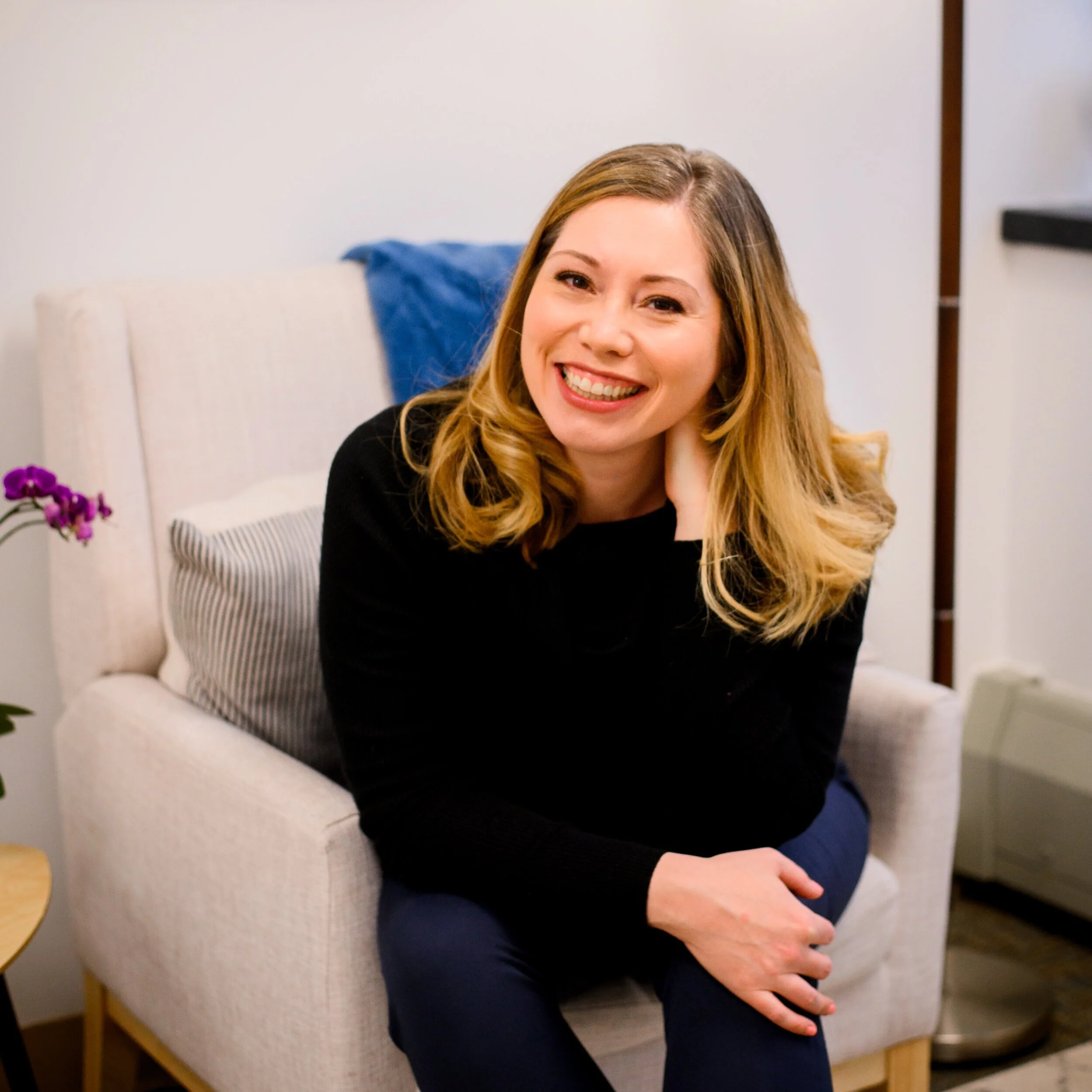 A woman with blonde hair wearing a black top and blue pants, smiling and sitting on a beige armchair in a cozy room.