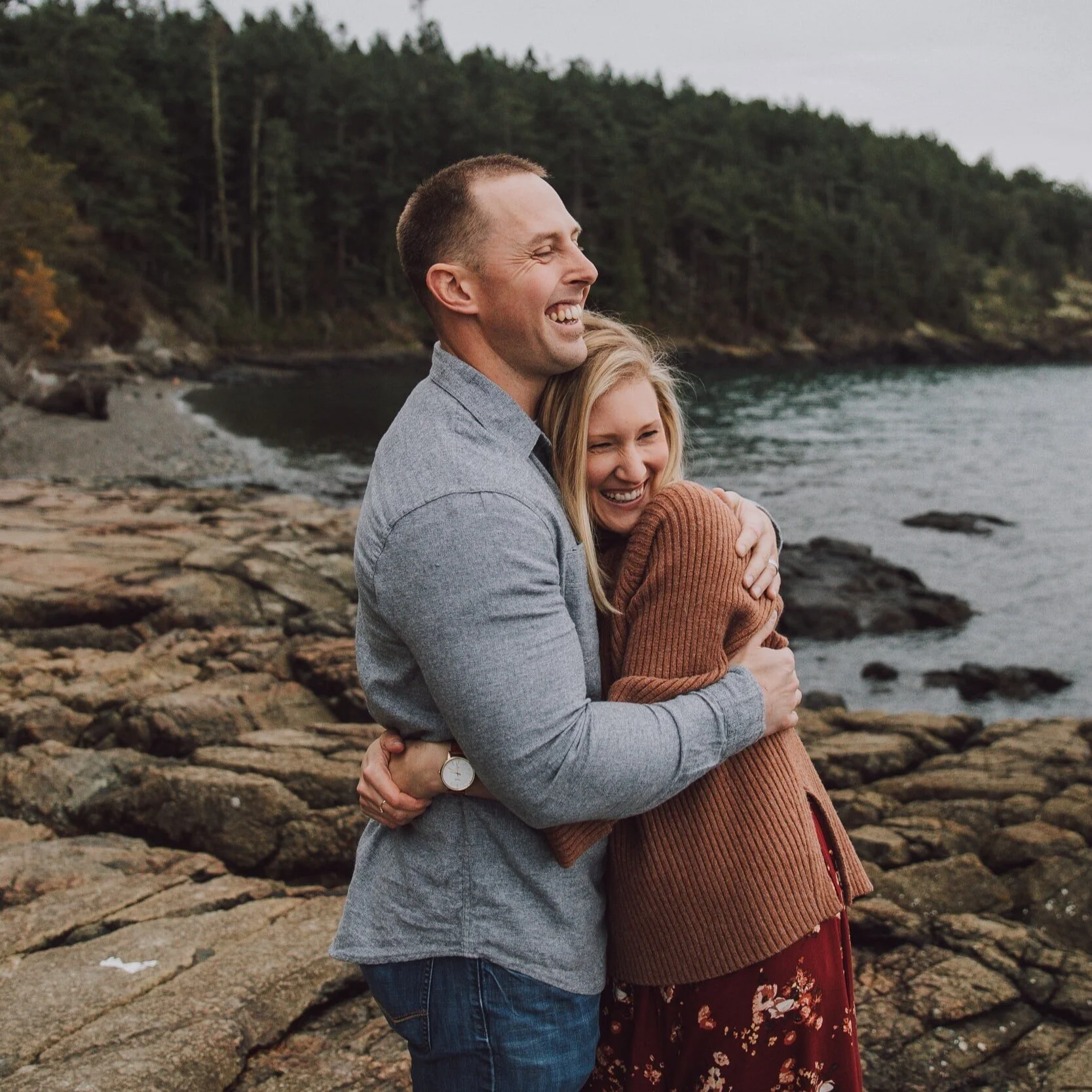 A happy couple hugging by the rocky shoreline with trees and water in the background.