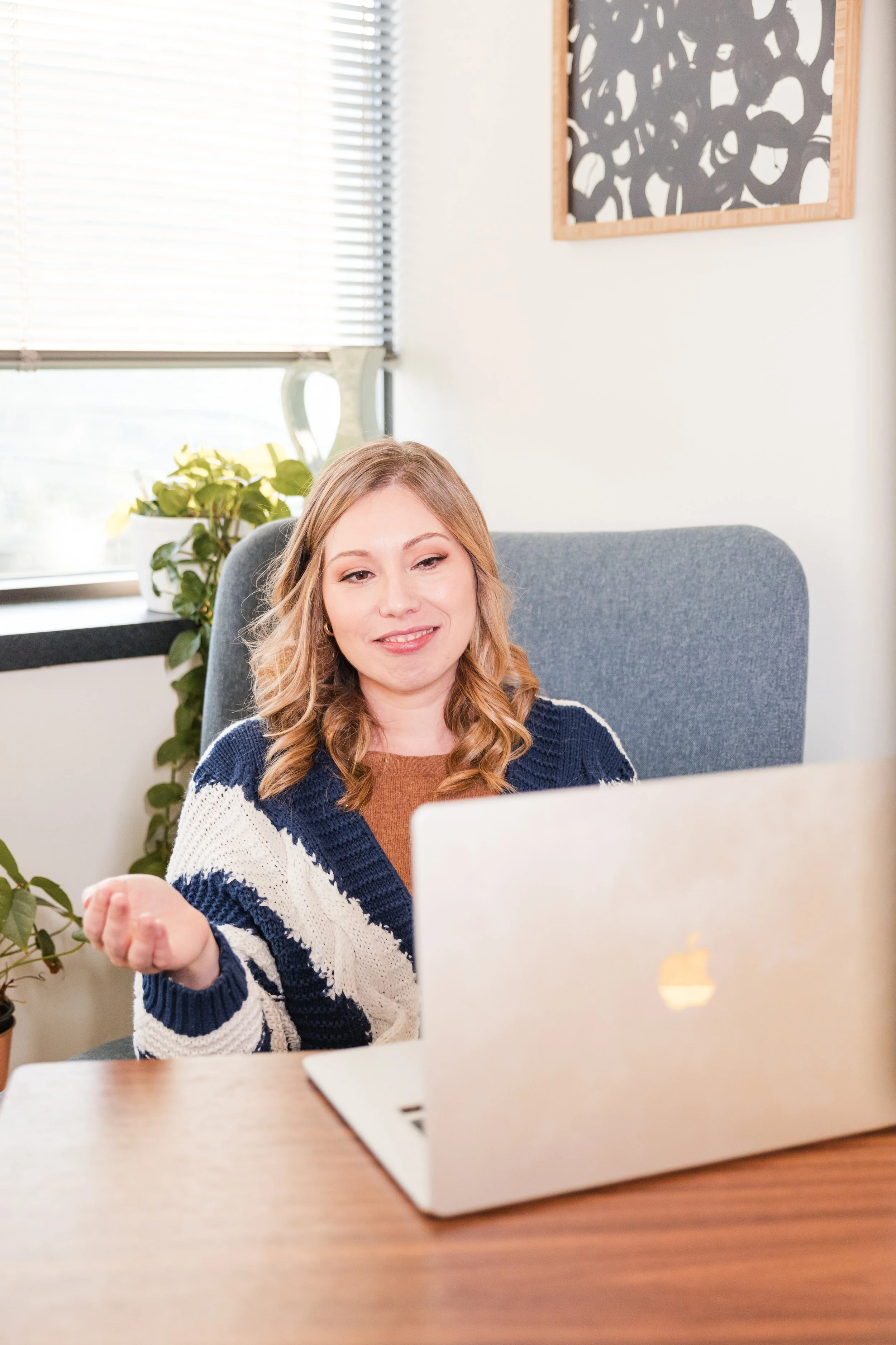 A woman with blonde hair sitting at a desk with a gray office chair, working on a silver MacBook laptop, in a bright office space with houseplants and abstract artwork on the wall.