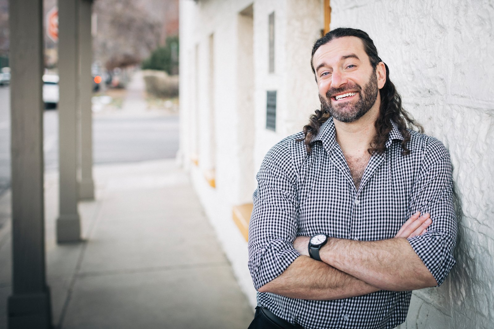 A man with long curly hair and a beard smiling, wearing a checkered shirt and a black watch, leaning against a white textured wall outdoors