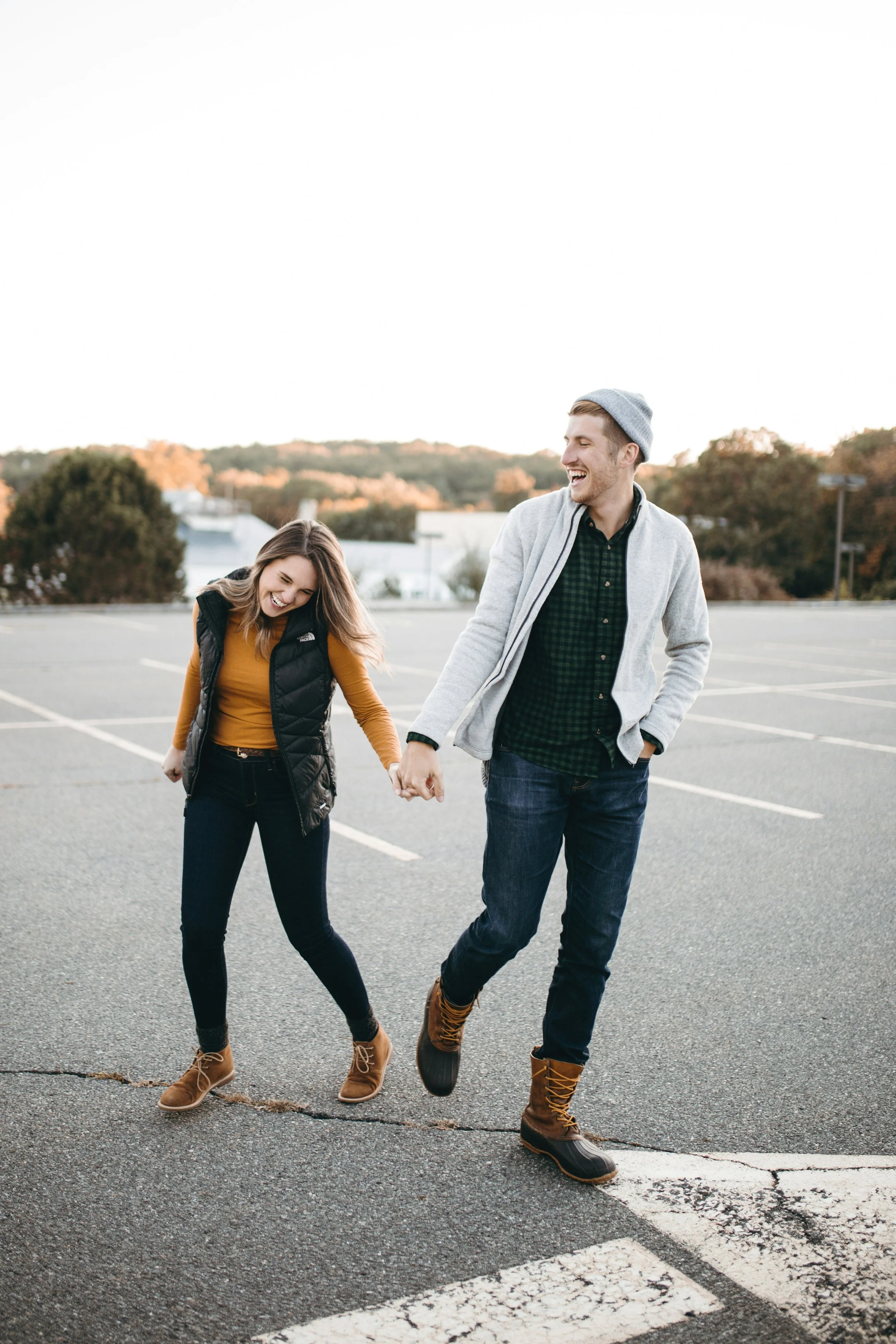 A young couple holding hands, laughing and walking in an empty parking lot during daytime, dressed casually with autumn scenery in the background.
