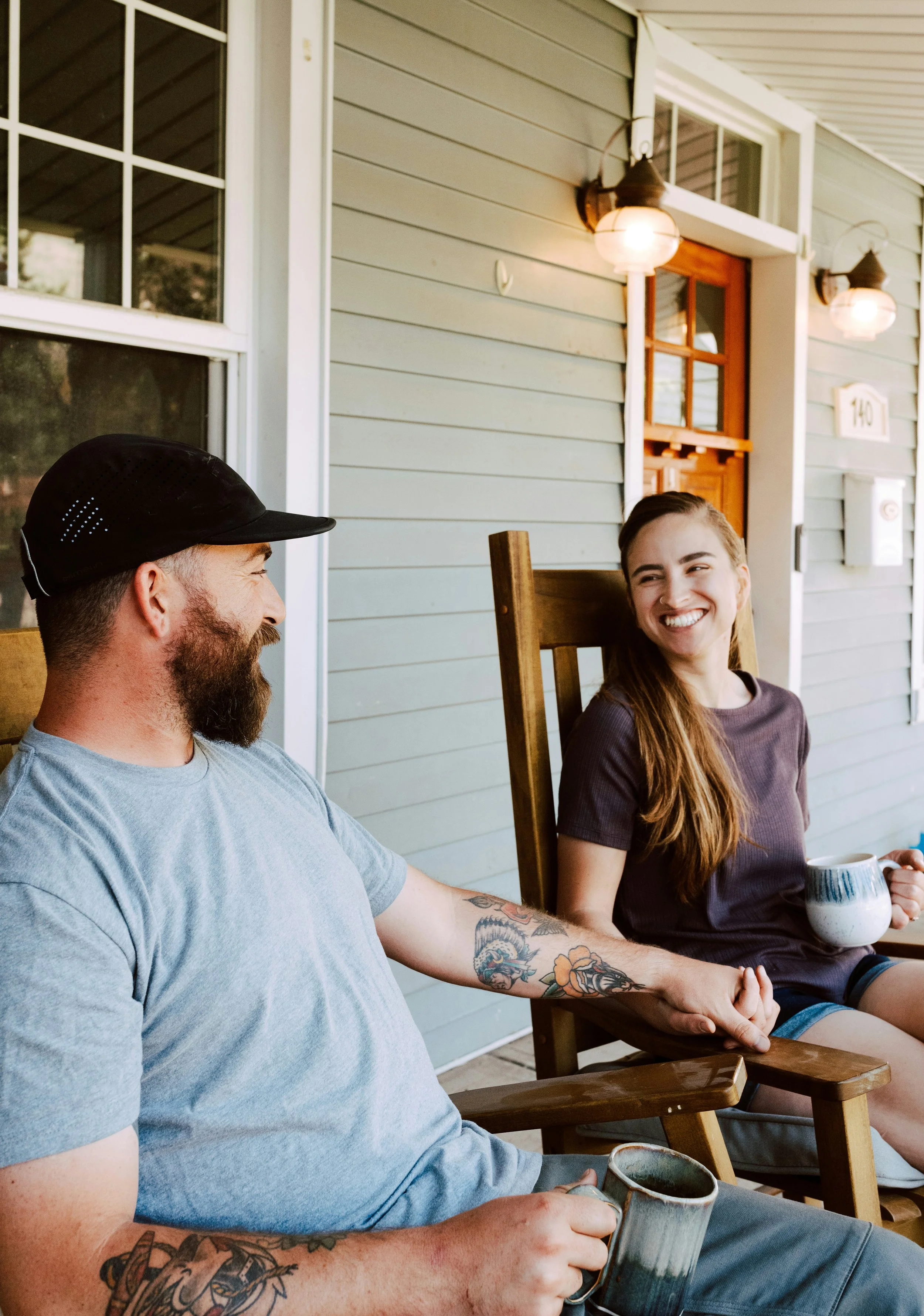A man and a woman sitting on a porch, holding hands and smiling at each other, with a coffee mug in the woman's hand.