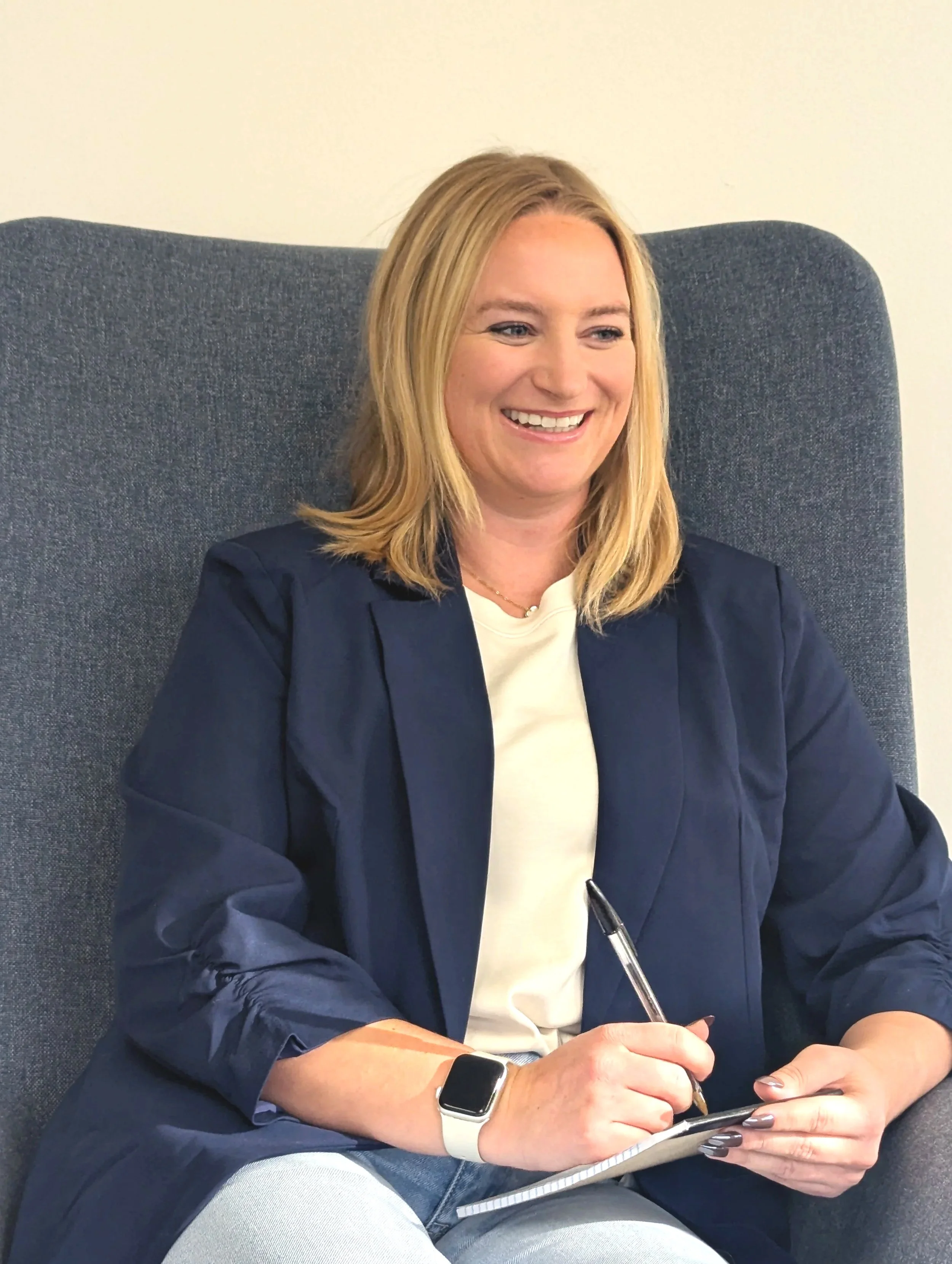 A woman with blonde hair sitting in a dark gray chair, smiling, wearing a navy blazer, a white shirt, and a smartwatch, holding a pen and a notepad.