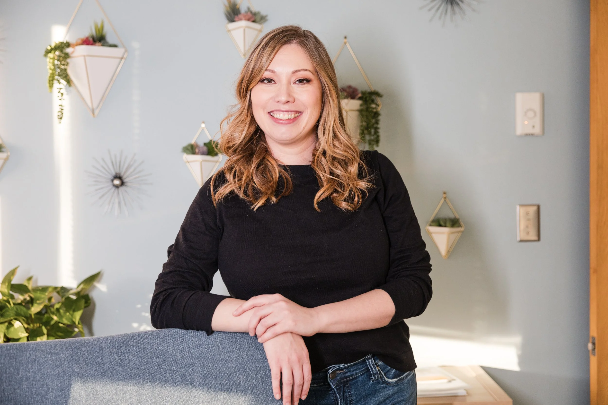 A smiling woman with wavy blonde hair, wearing a black long-sleeve shirt, standing indoors with her hands resting on a blue chair. The background has light blue walls decorated with geometric planters and plants.