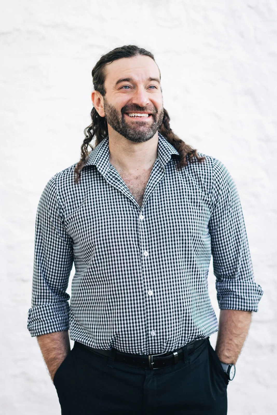 A smiling man with long curly hair and a beard, wearing a black and white checkered shirt, standing with his hands in his pockets against a white background.