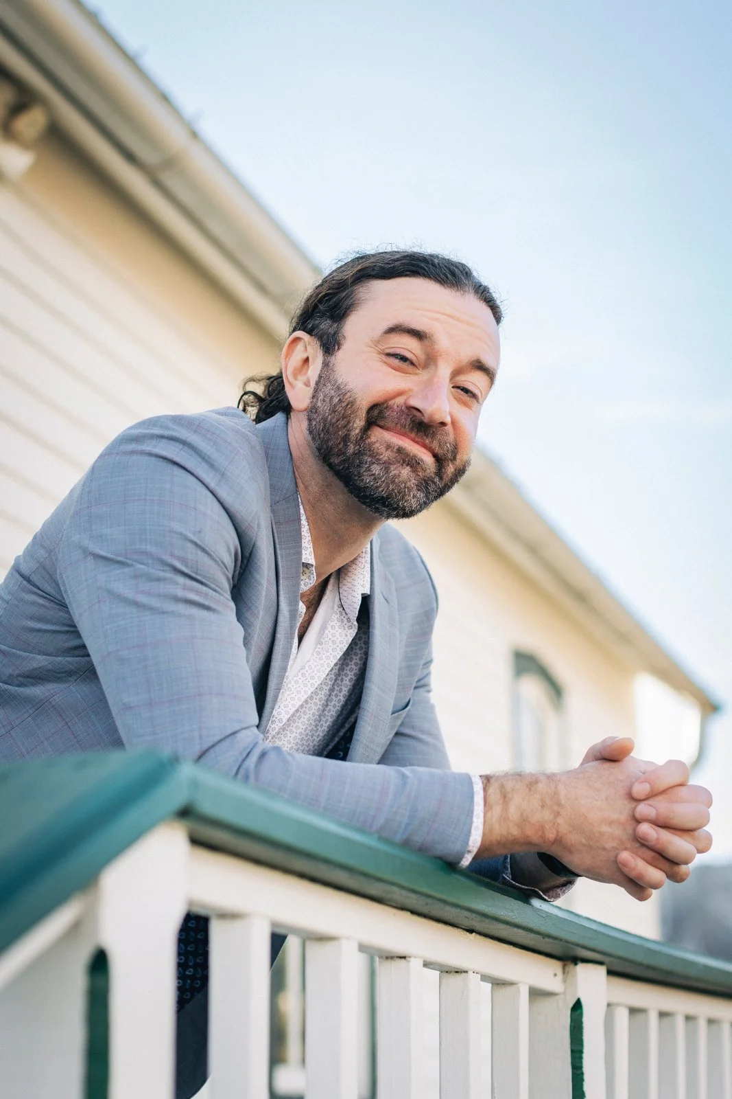 A man with dark hair and a beard, wearing a light gray suit, smiling and leaning on a porch railing outdoors.
