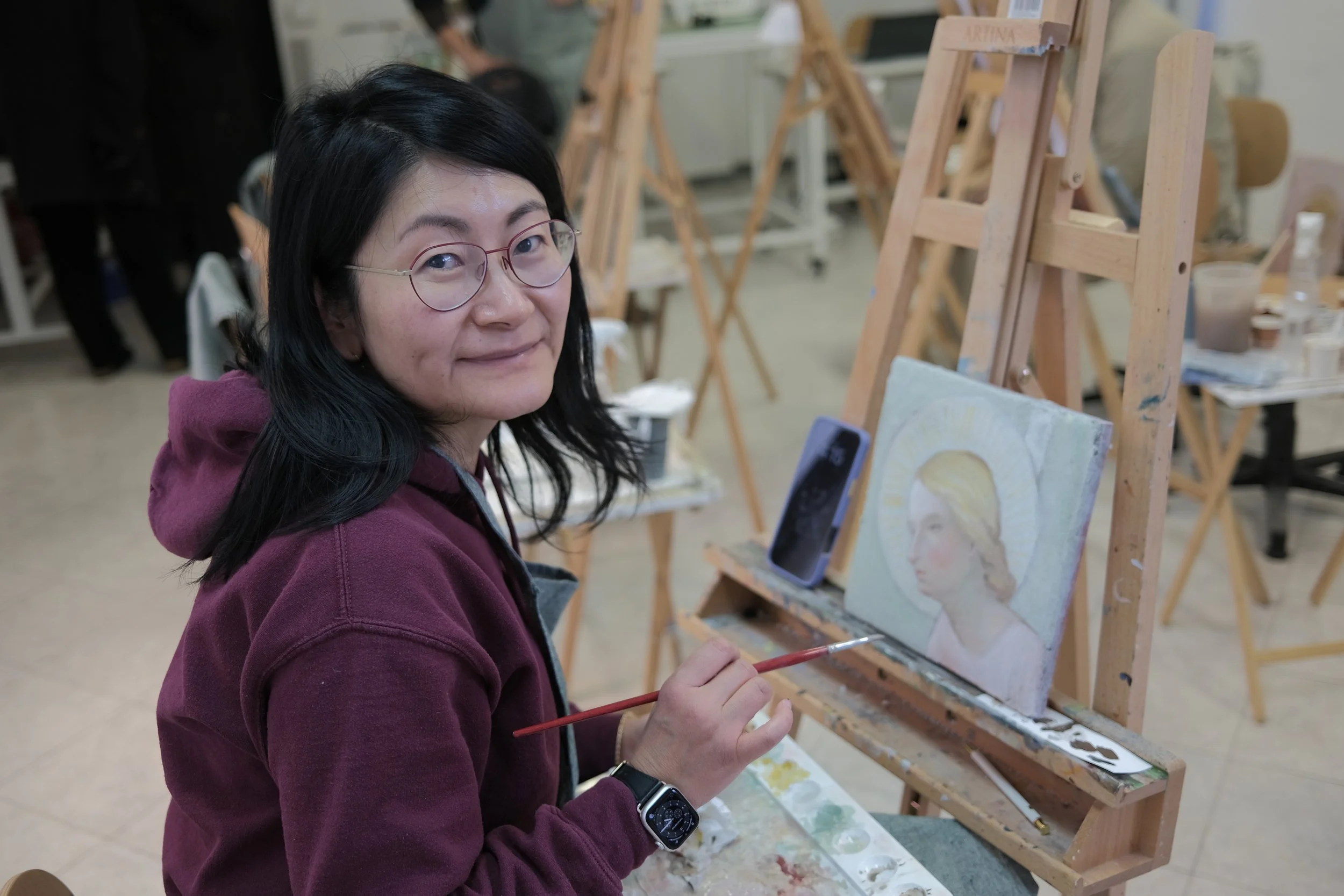 A woman with black hair and glasses, wearing a maroon hoodie, is painting a portrait on a canvas in an art studio. She is holding a paintbrush and is seated in front of an easel with her artwork.