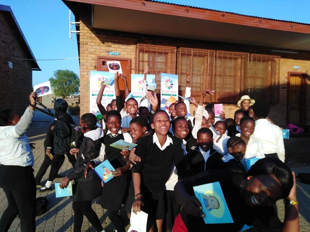A group of students outside a school building, smiling and holding up colorful pamphlets and books, celebrating together on a sunny day.