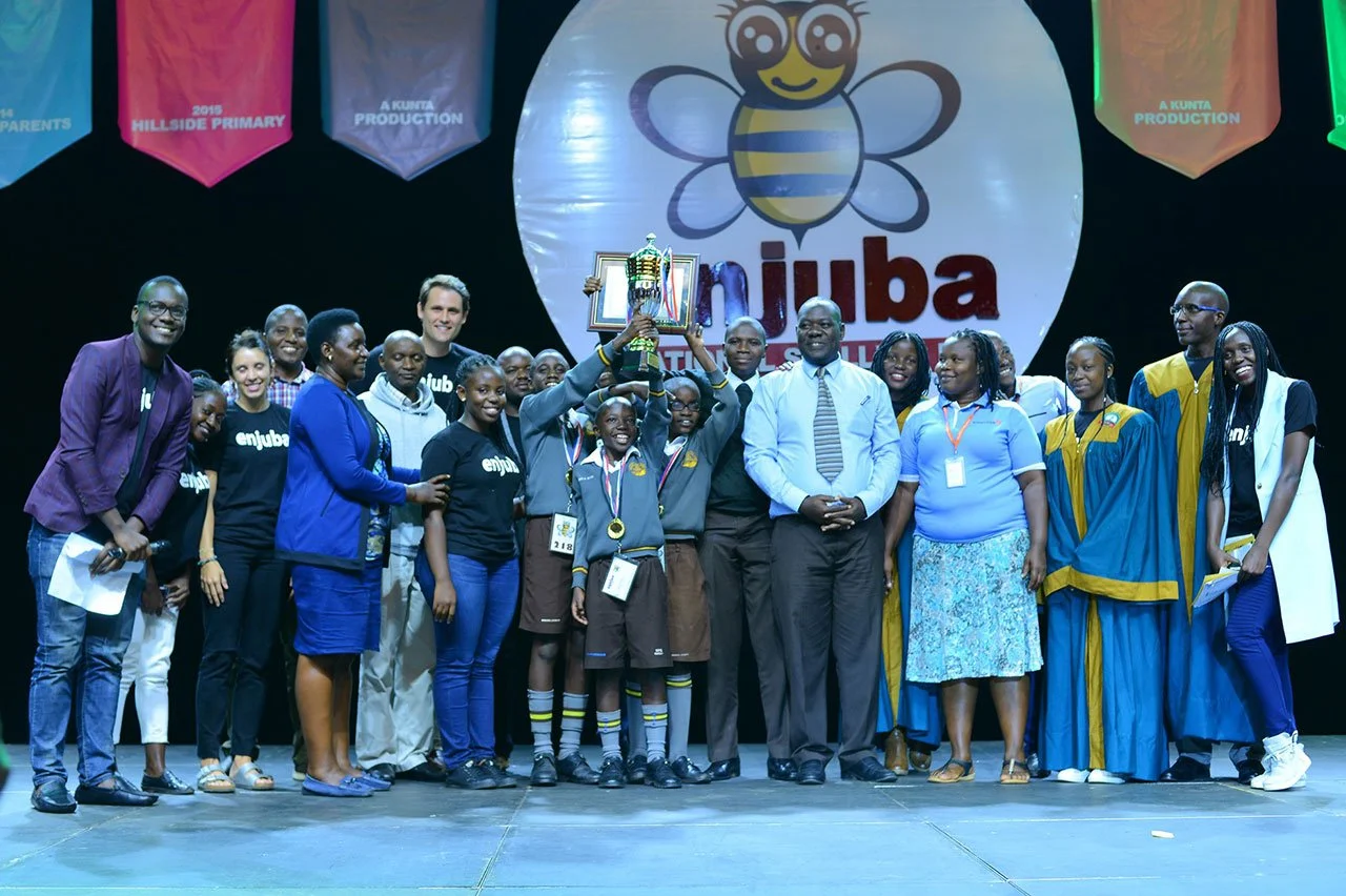 A group of children, teachers, and officials on stage holding a trophy after a competition, with banners and a large logo featuring a smiling bee in the background.