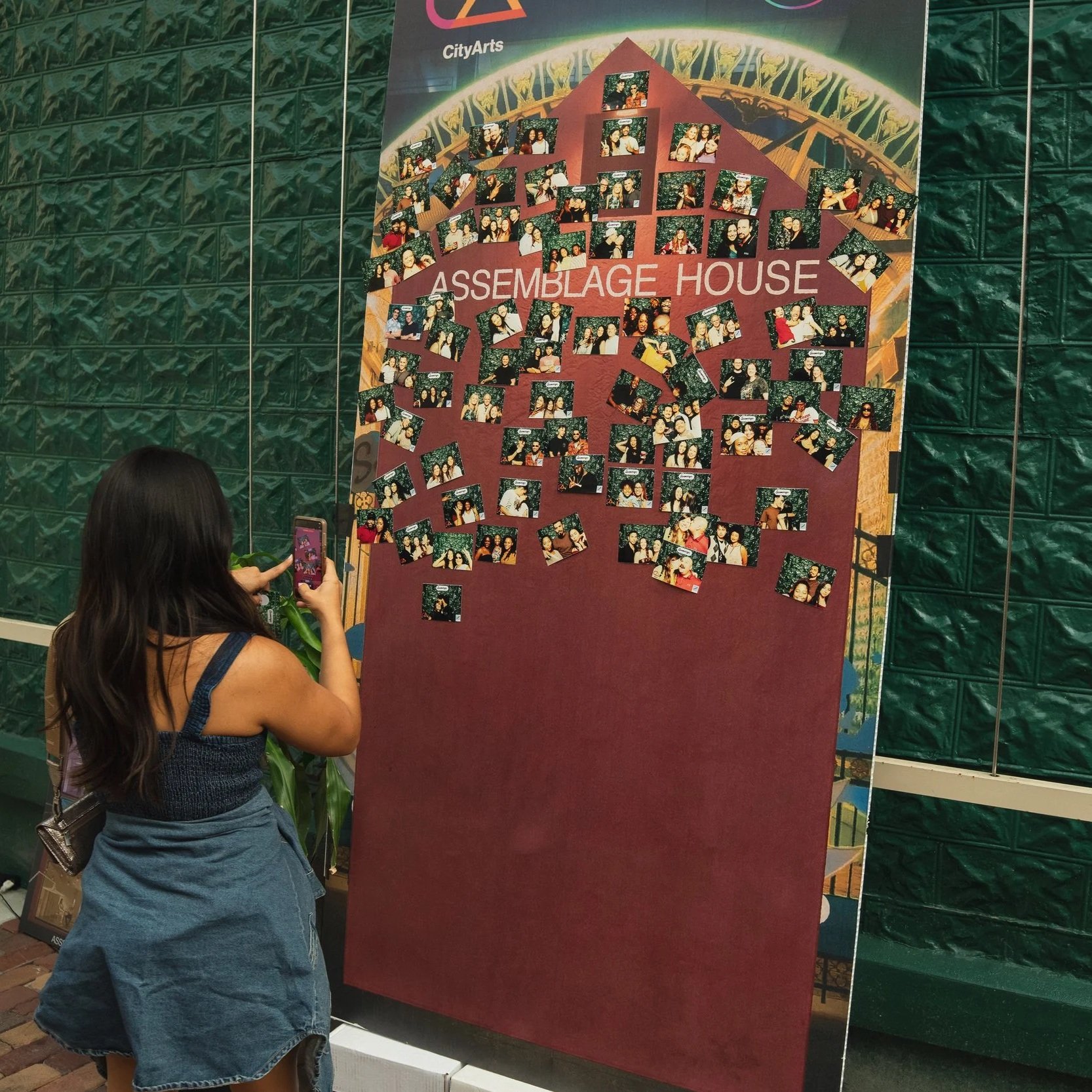 A photo of a woman taking a picture of a photo wall after placing her printed picture on to it.
