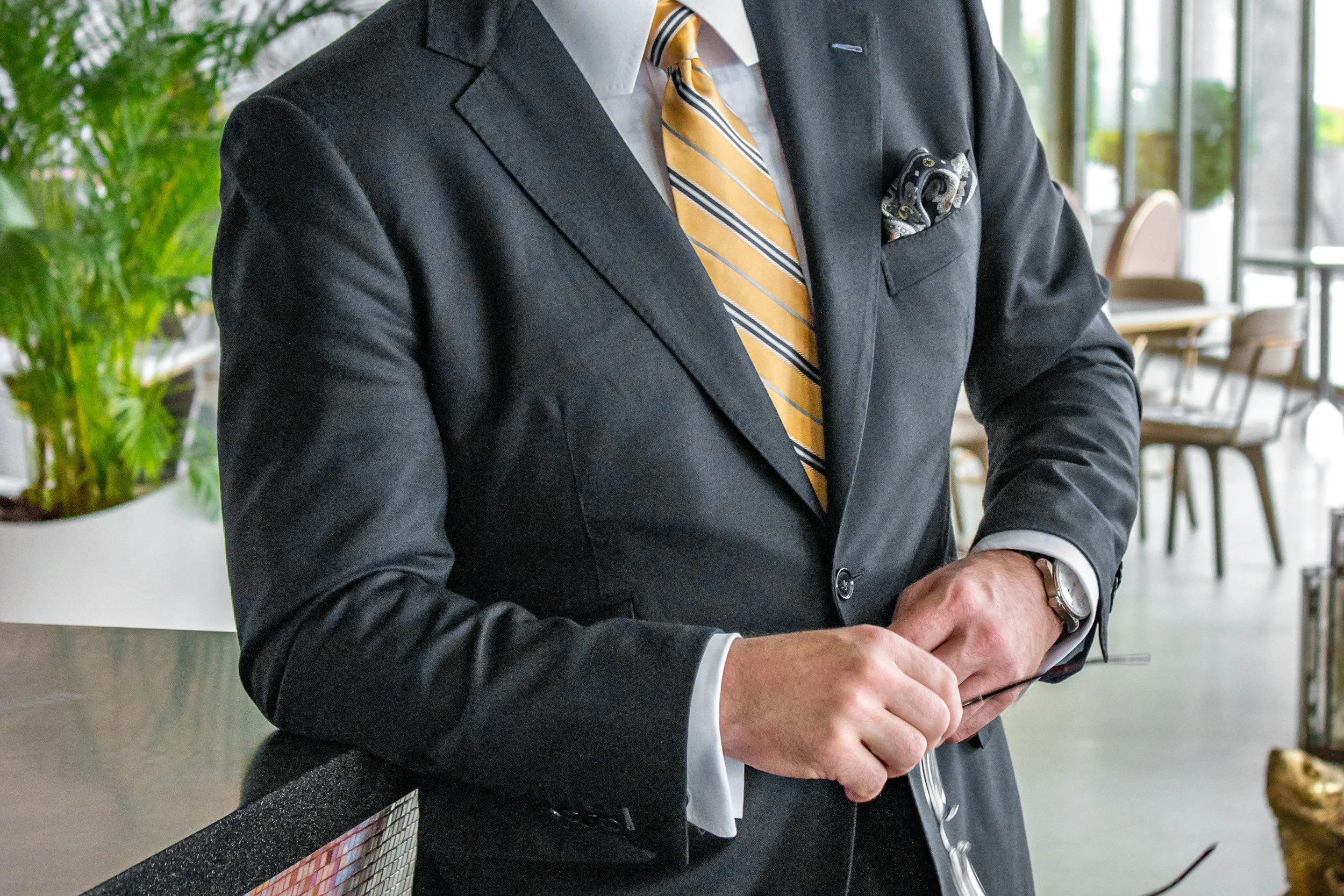 A man in a dark suit with a yellow striped tie, holding a smartphone in a modern office or cafe setting with chairs, tables, and greenery in the background.