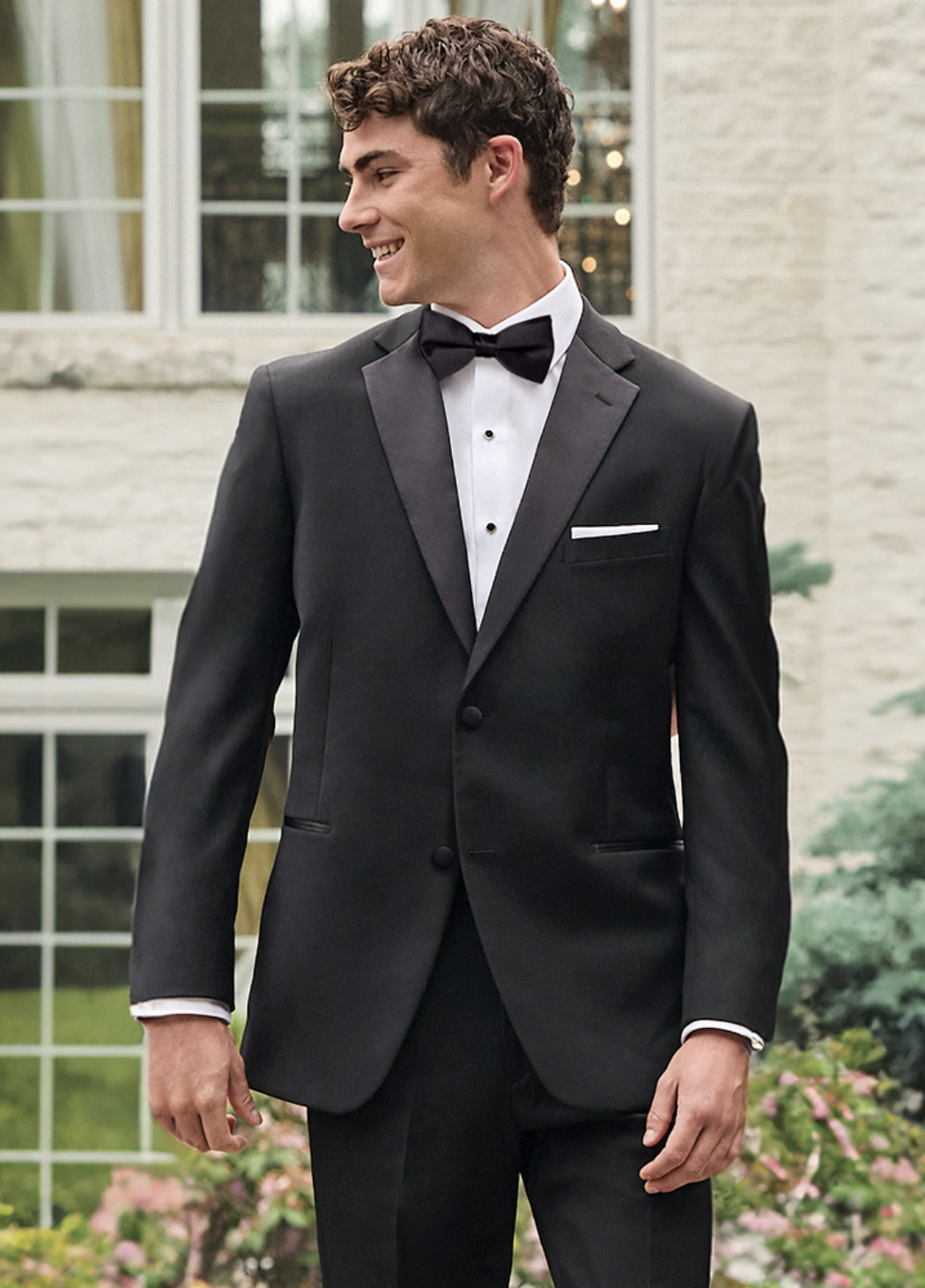 A young man wearing a black tuxedo, white dress shirt, and black bow tie, standing outside near a large window and greenery, smiling and looking to his left.