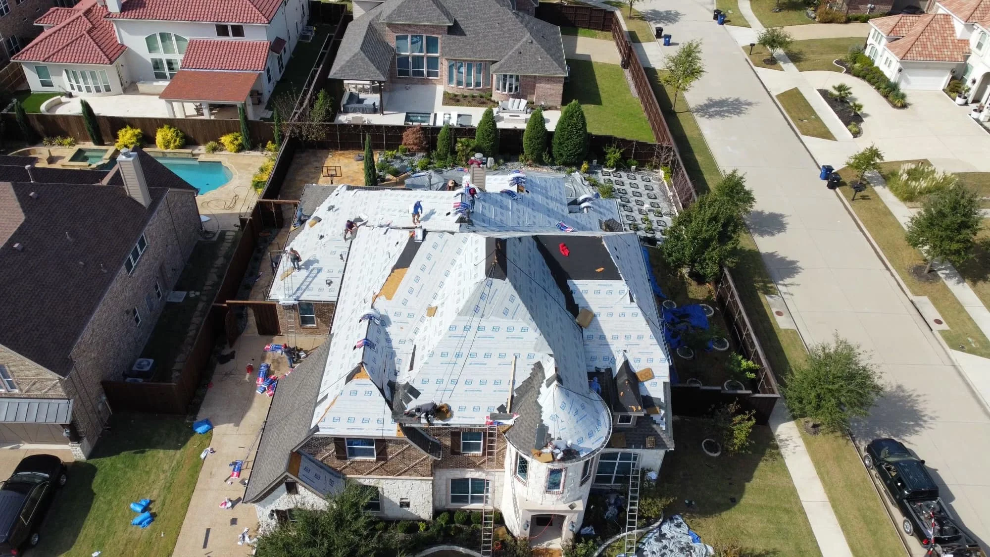 Aerial view of a residential neighborhood with roof construction. Workers are installing protective roofing material on a large house. Nearby houses have completed roofs and swimming pools. The street is visible with trees and parked cars.