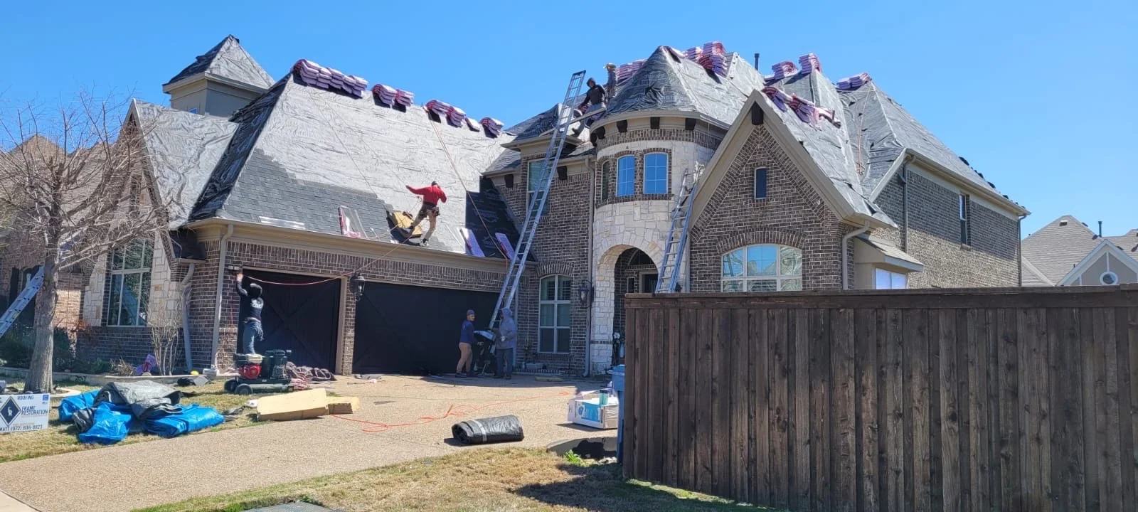 Workers on a steep roof of a large brick house performing roof repairs; ladders and roofing materials visible.
