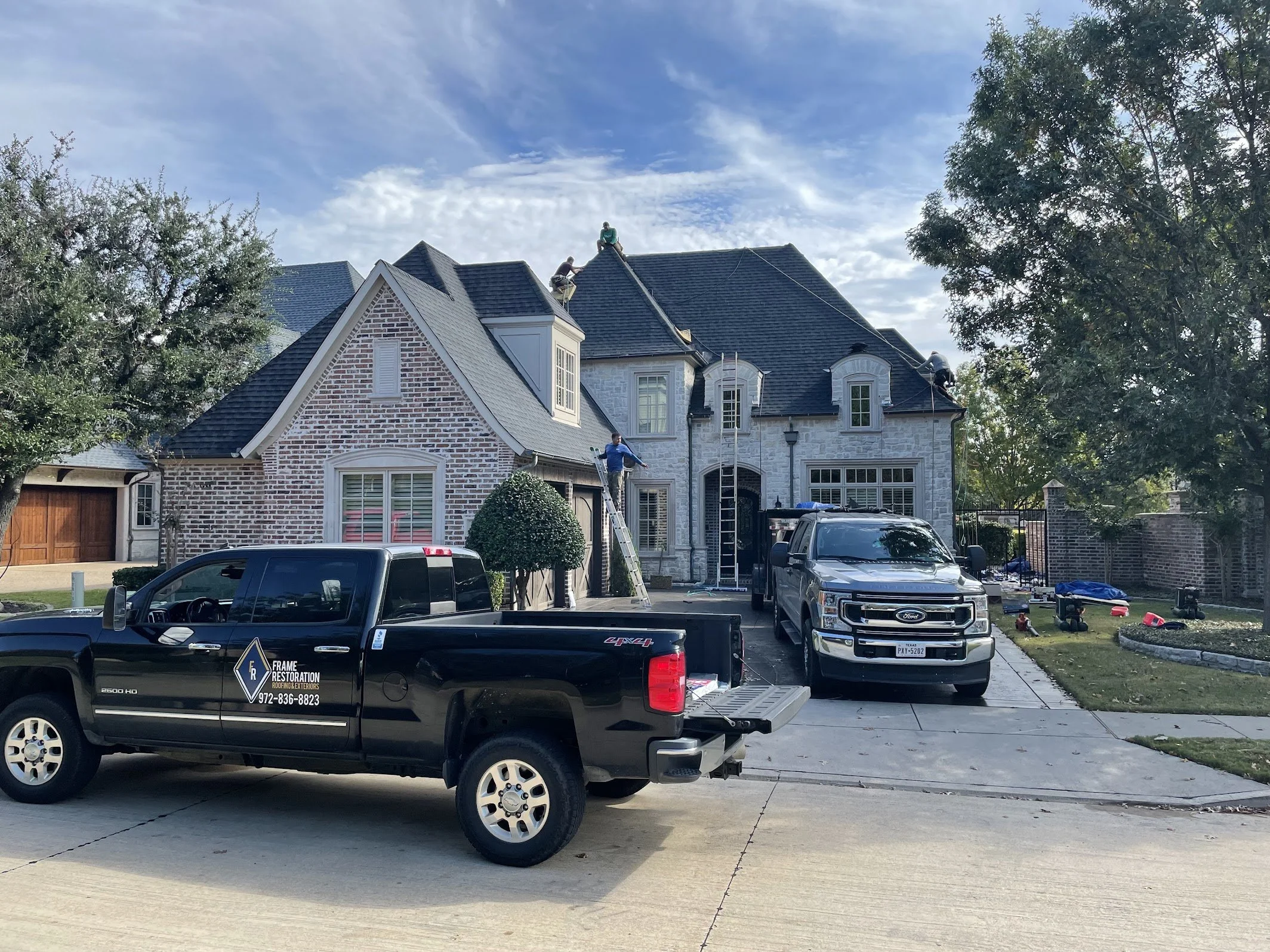 Brick house with workers on roof, two pickup trucks parked in front, and trees surrounding the area.