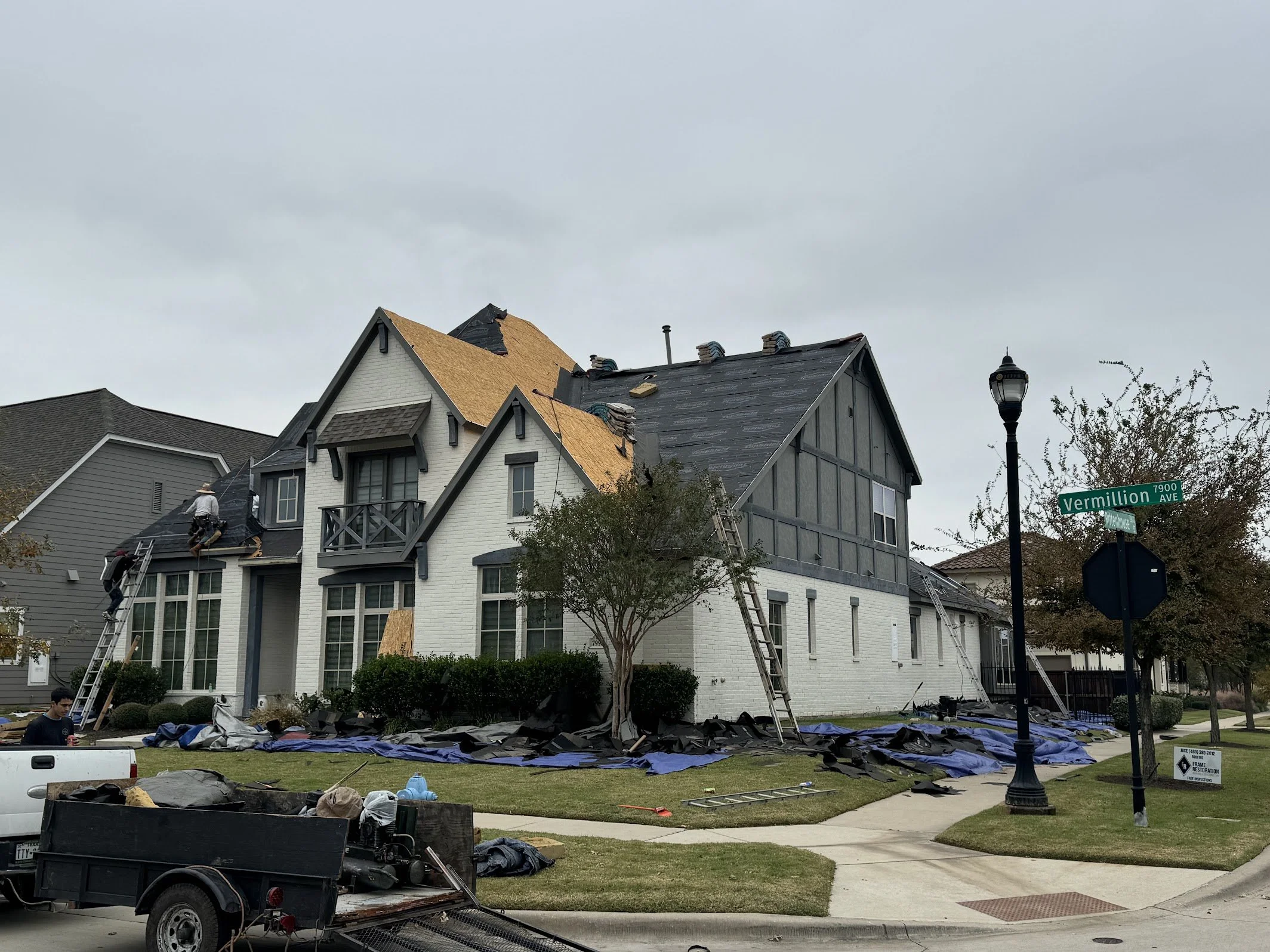 Construction workers repairing roof of a white house on a corner lot at Vermillion Avenue, with tools, ladders, and roofing materials on the lawn and street.