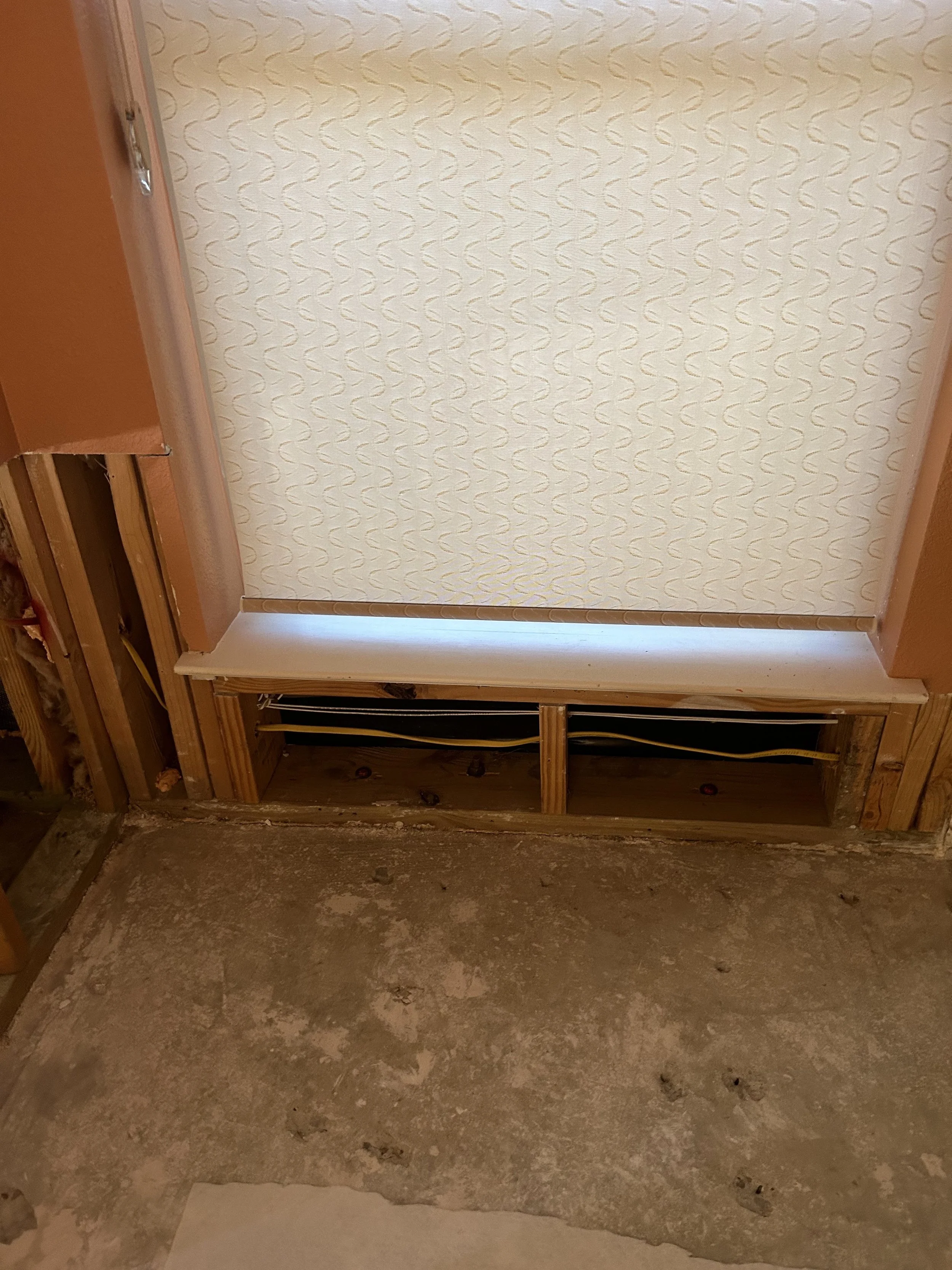Interior view of a partially unfinished wall next to a window with a patterned roller blind, showing exposed wooden framing and electrical wires.