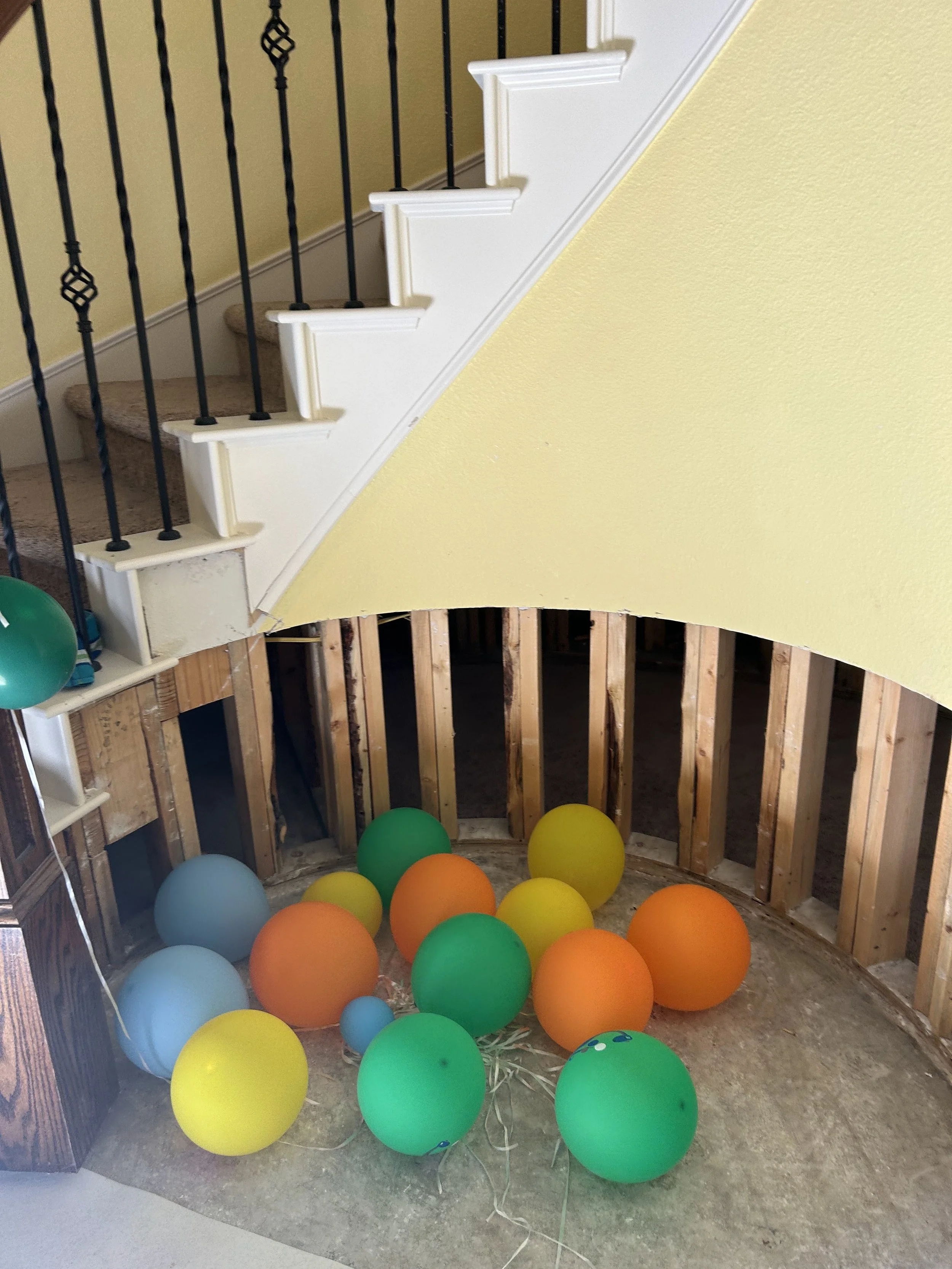 Colorful balloons on the floor underneath a staircase with missing drywall, revealing wooden studs.