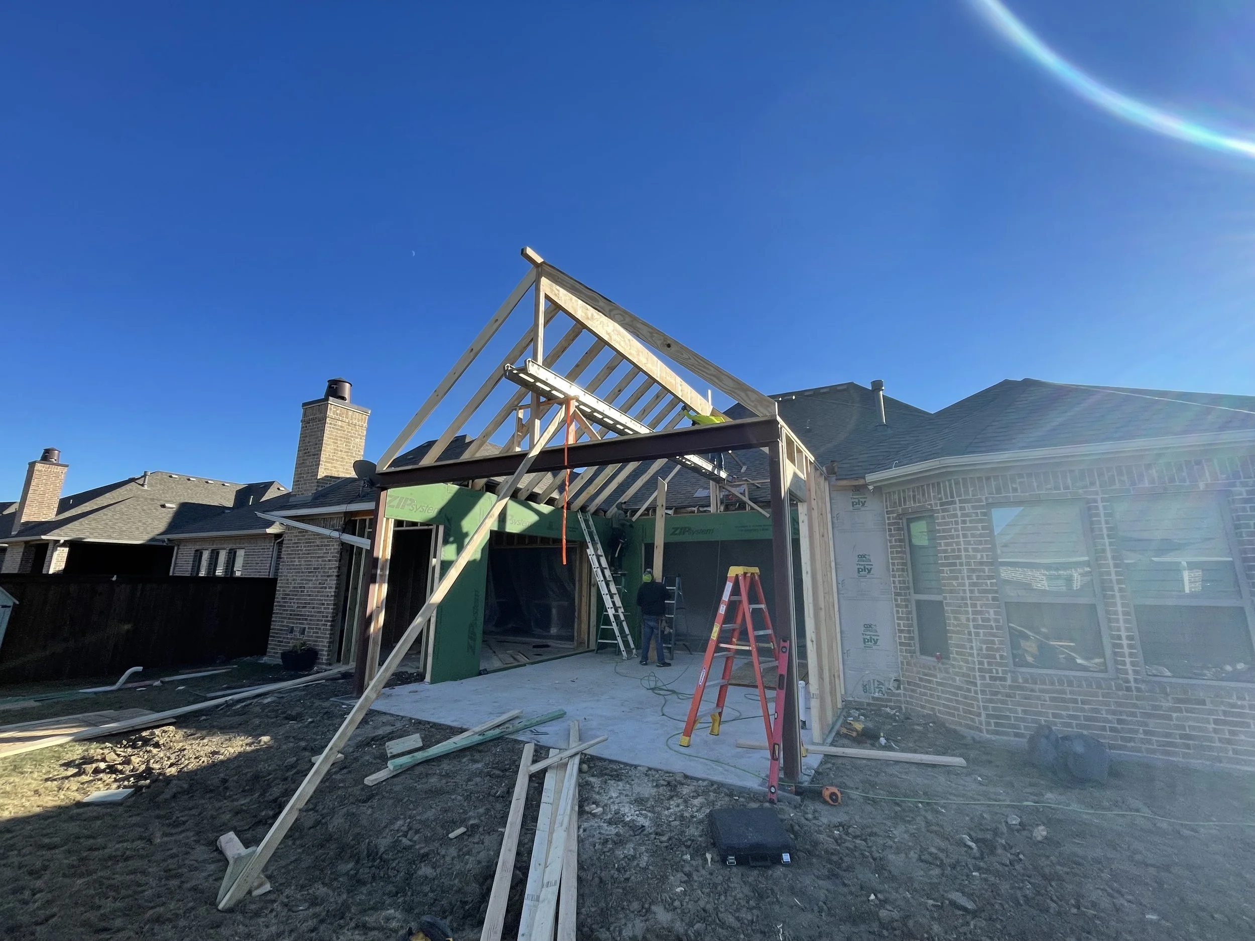Home construction site with partially built structure, wooden framework visible, and construction materials scattered. Clear blue sky overhead.
