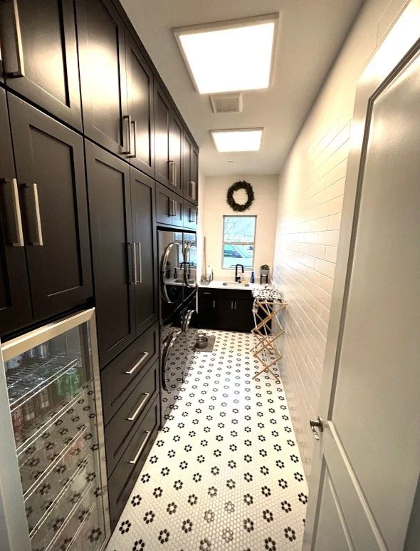 Modern laundry room with dark cabinets, a washing machine, dryer, and countertop sink. Features a mini fridge and drying rack on black-and-white tiled floor, with overhead lights and a window.