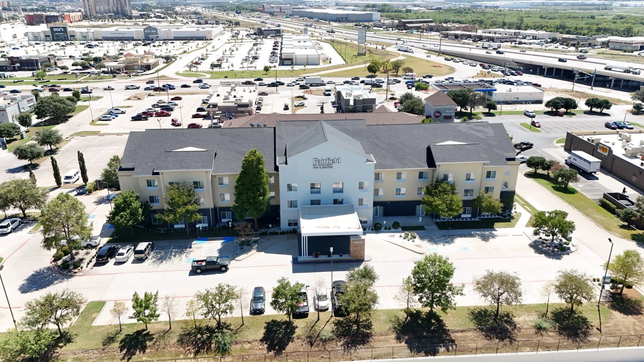 Aerial view of a hotel with parking lot, surrounded by trees and buildings, near a highway in an urban area.