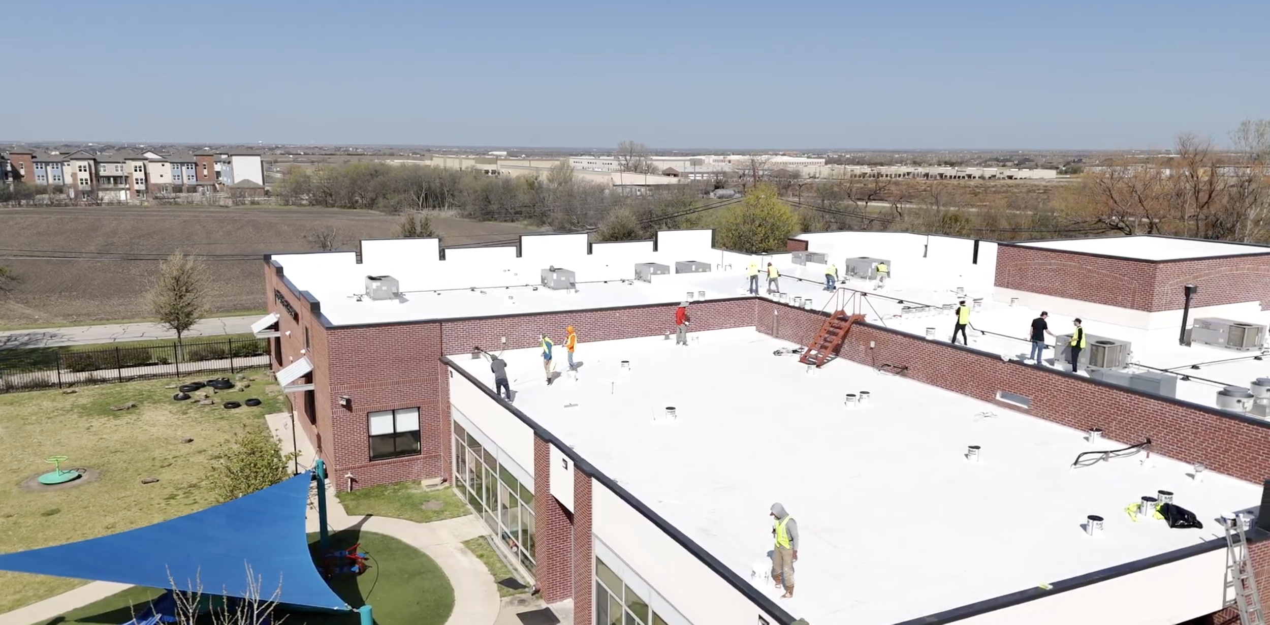 Aerial view of a building with a white rooftop where several construction workers are present. The building has a red brick exterior and is surrounded by open fields and trees. A playground structure with a blue canopy is visible in the yard.