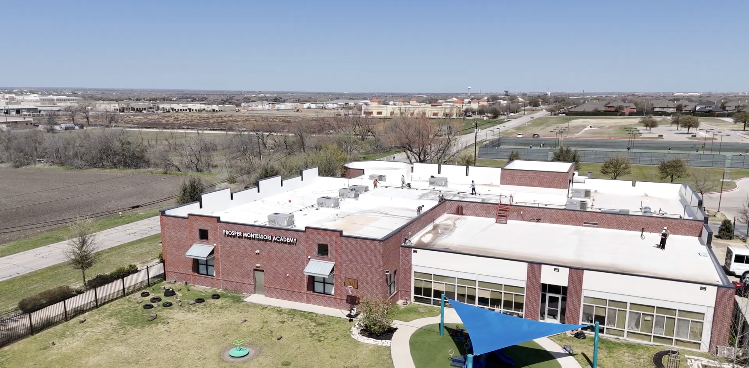 Aerial view of Prosper Montessori Academy, a red brick building with a playground featuring a blue sun shade. The surrounding area includes roads, tennis courts, and open fields, with several people visible on the building's roof.