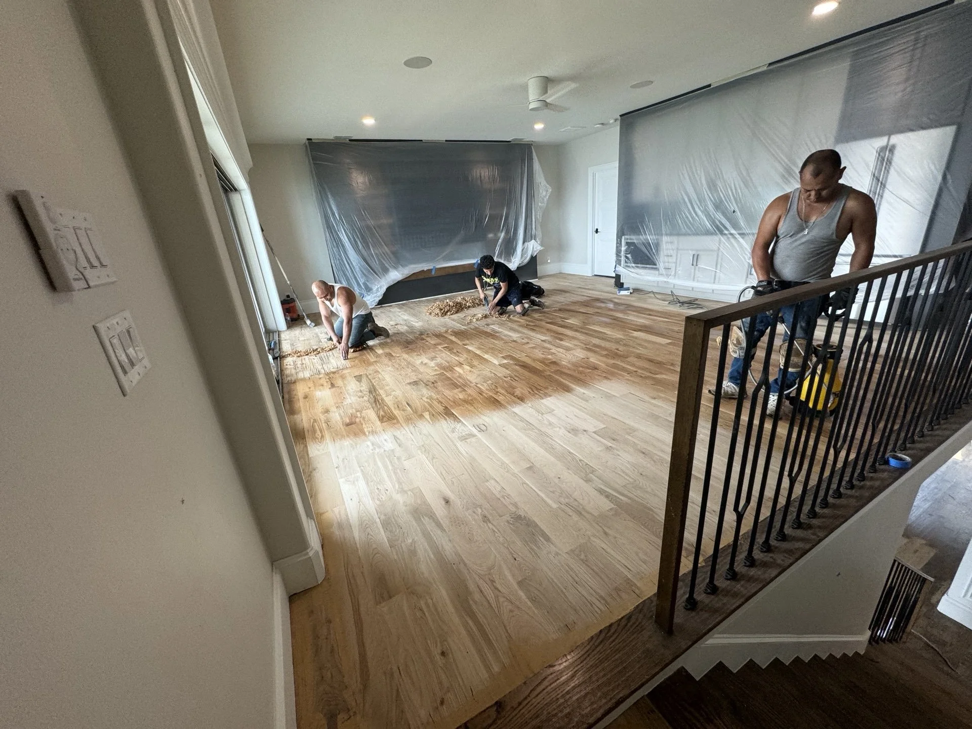 Workers installing hardwood flooring in a room with covered walls and tools.