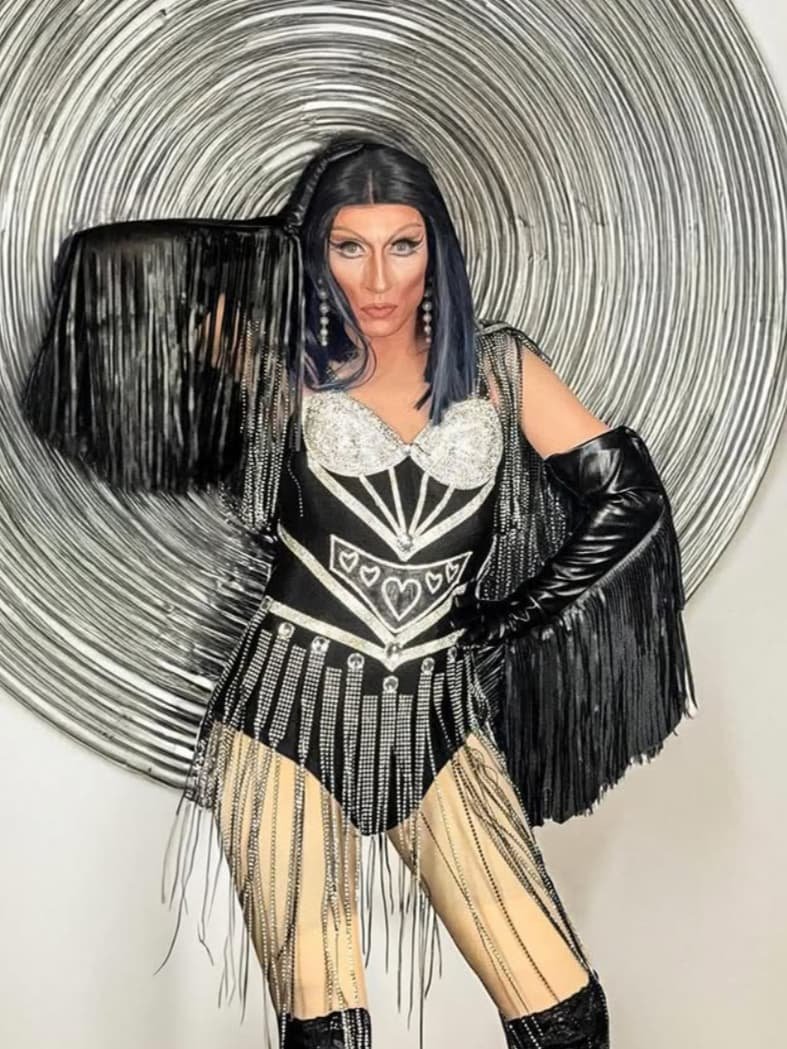 Drag-Künstlerin Liz Queera posiert in einem schwarzen Fransen-Body mit einem silber-glitzernden Bustier vor einem großen, spiralförmigen Kunstwerk.