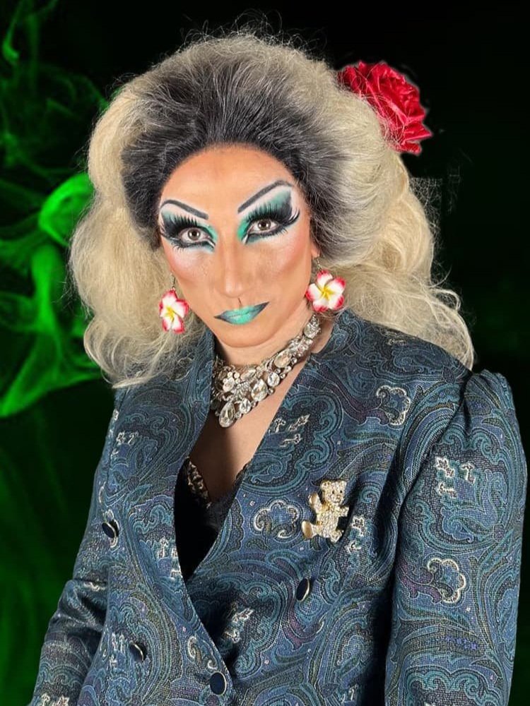 Studioporträt der Drag Queen Liz Queera mit auffälligem grünen Lidschatten und Lippenstift, gekleidet in einem blauen Paisley-Blazer mit einer Teddybär-Brosche.