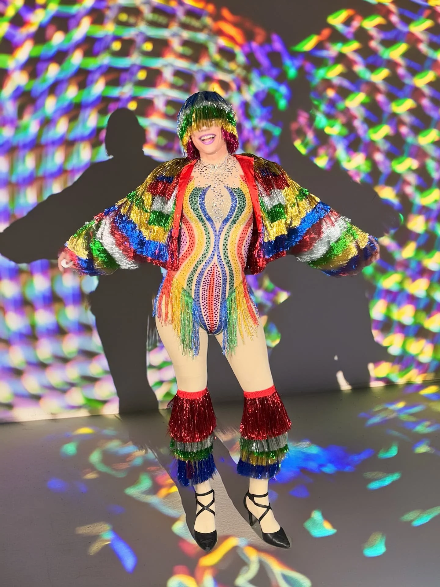 Drag-Künstlerin Liz Queera in einem bunten, glitzernden Karnevals-Outfit mit Fransen-Jacke und Regenbogen-Body.