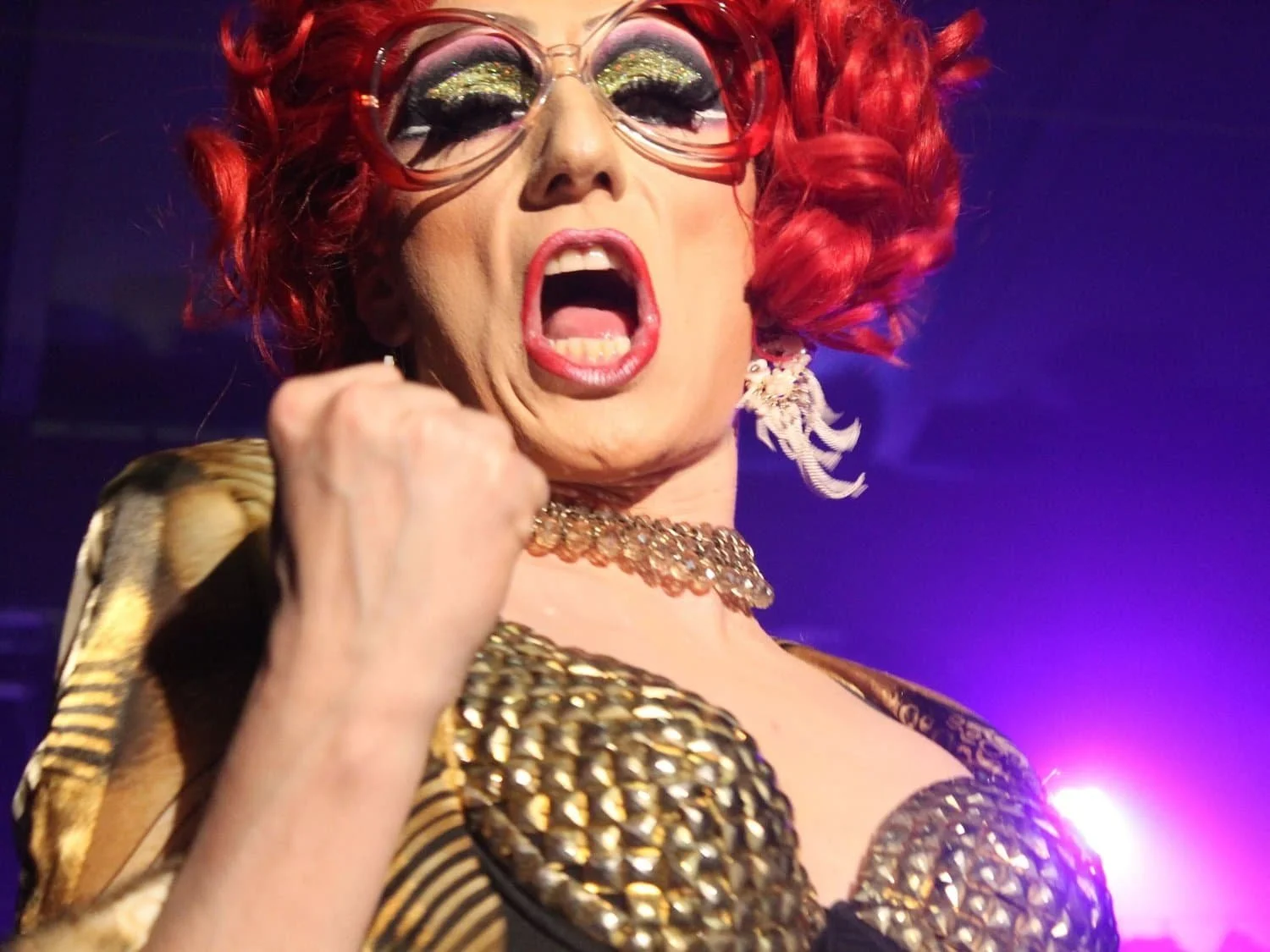 Nahaufnahme von Liz Queera in einem goldenen Bustier, die mit geballter Faust und intensivem Ausdruck auf der Bühne singt.