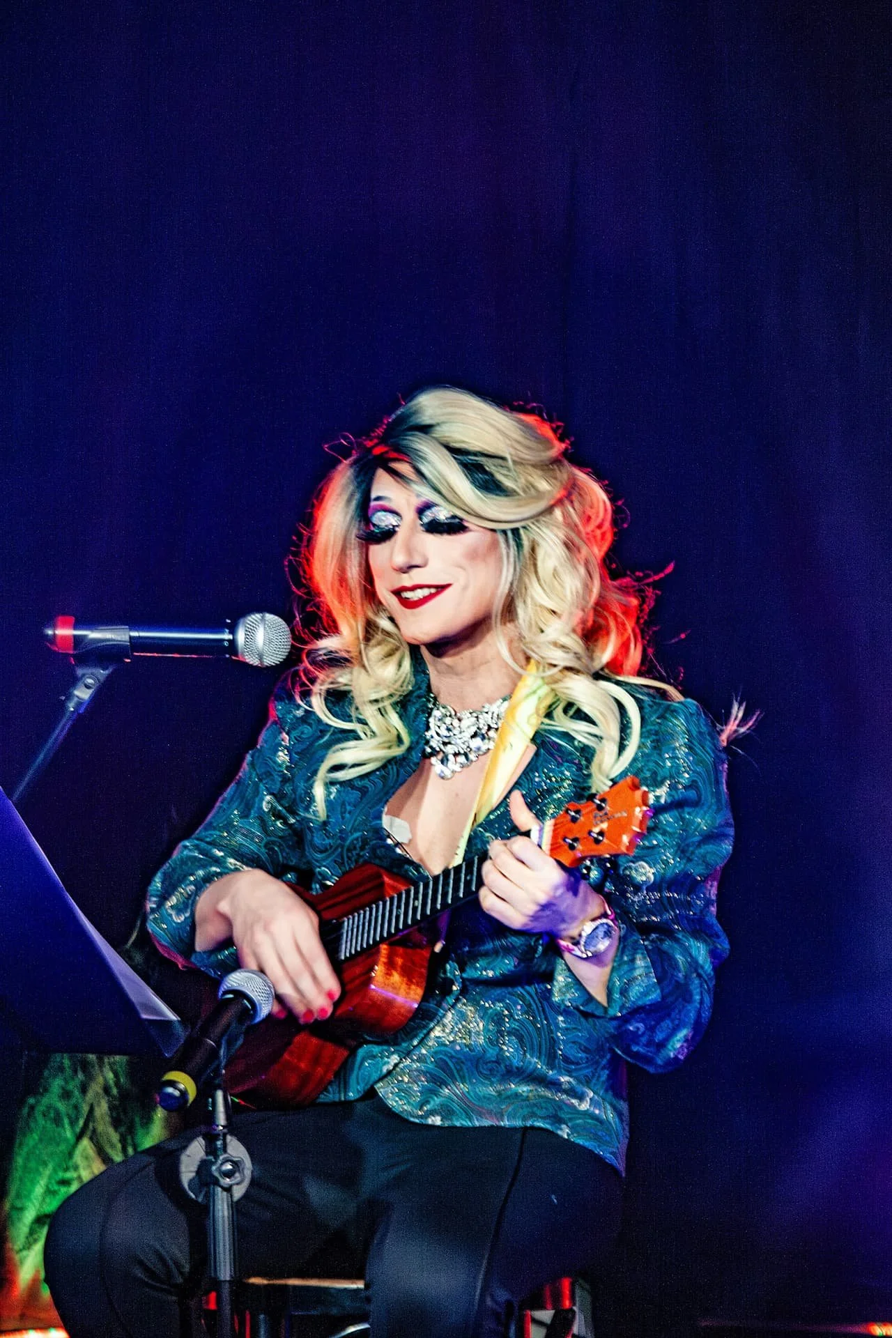 Drag-Künstlerin Liz Queera sitzt auf der Bühne, singt ins Mikrofon und spielt dabei auf ihrer Ukulele.