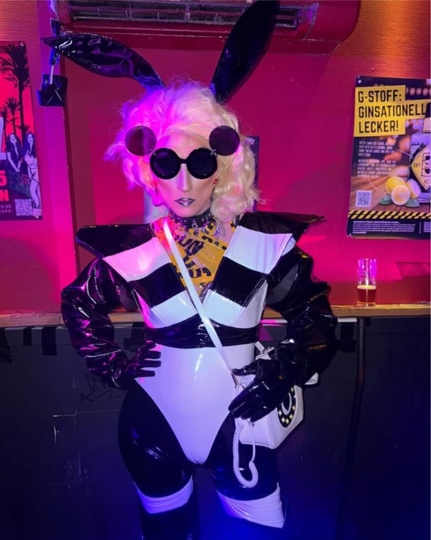 Liz Queera in einem futuristischen schwarz-weißen Vinyl-Body mit großen Hasenohren und einer Handtasche in Telefon-Optik.