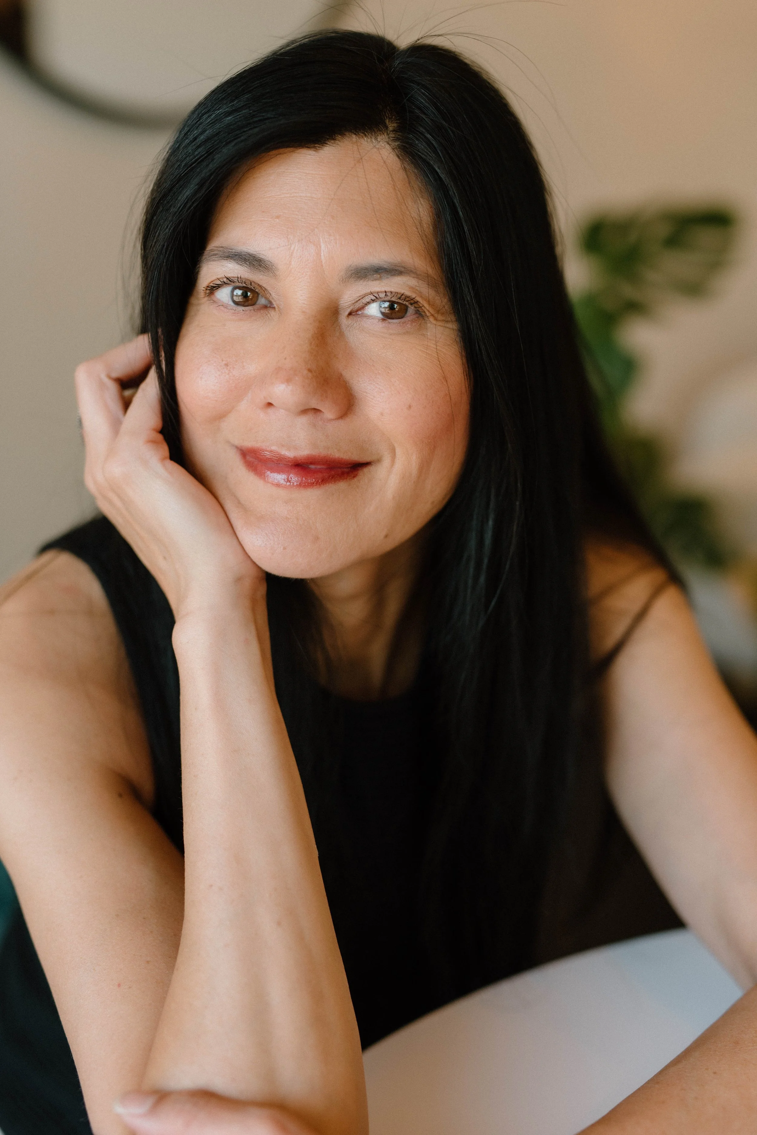 A woman with long black hair, wearing a black sleeveless top, sitting indoors, resting her face on her hand, smiling softly at the camera.