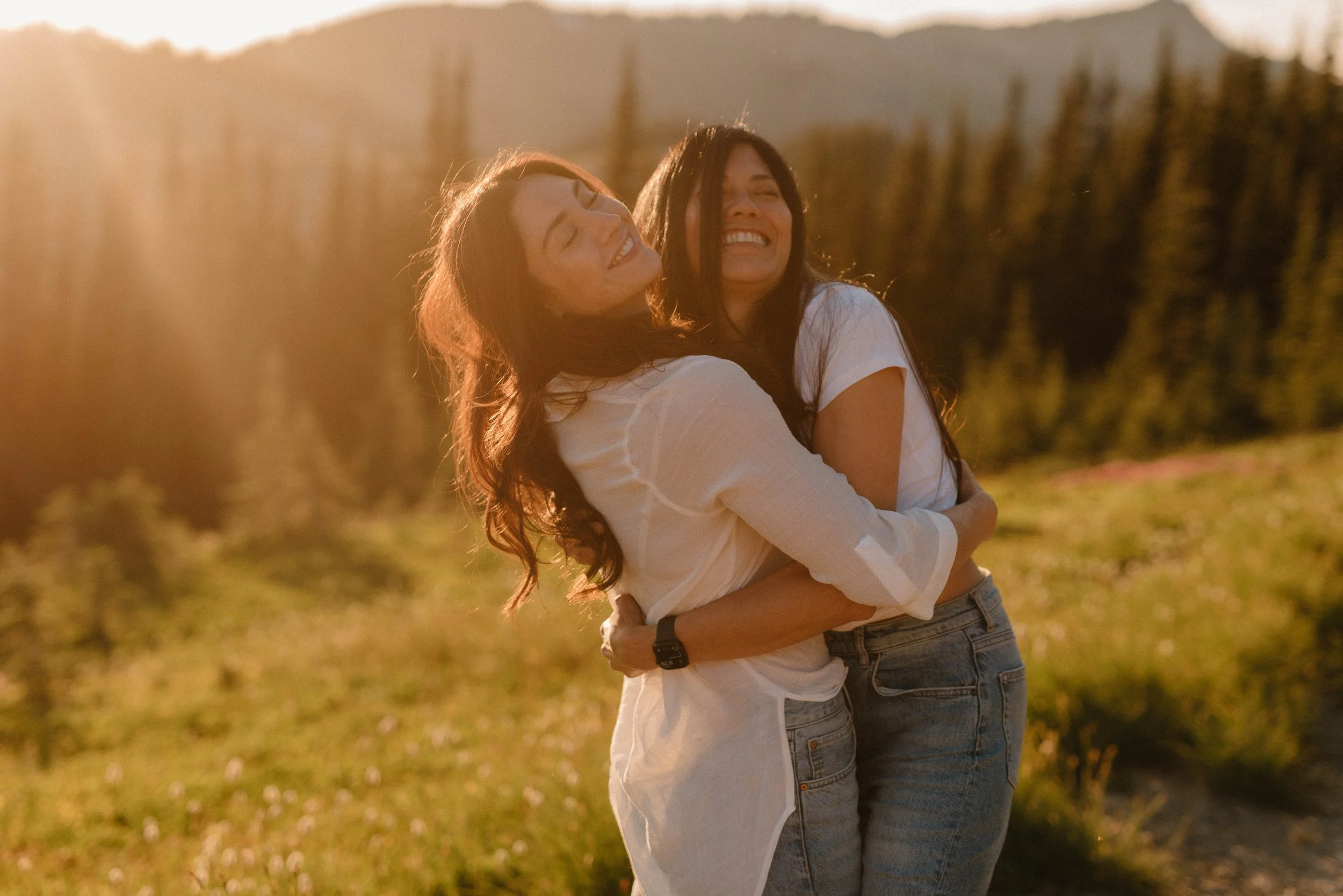 Two women hugging and smiling outdoors during sunset with a field and forest in the background.