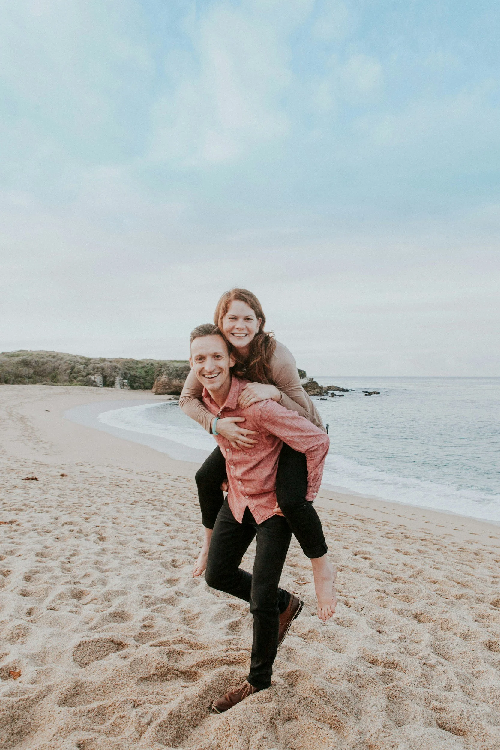 A couple on a sandy beach, with the woman on the man's back, smiling and showing a playful piggyback pose against a background of the ocean, rocky shoreline, and a partly cloudy sky.