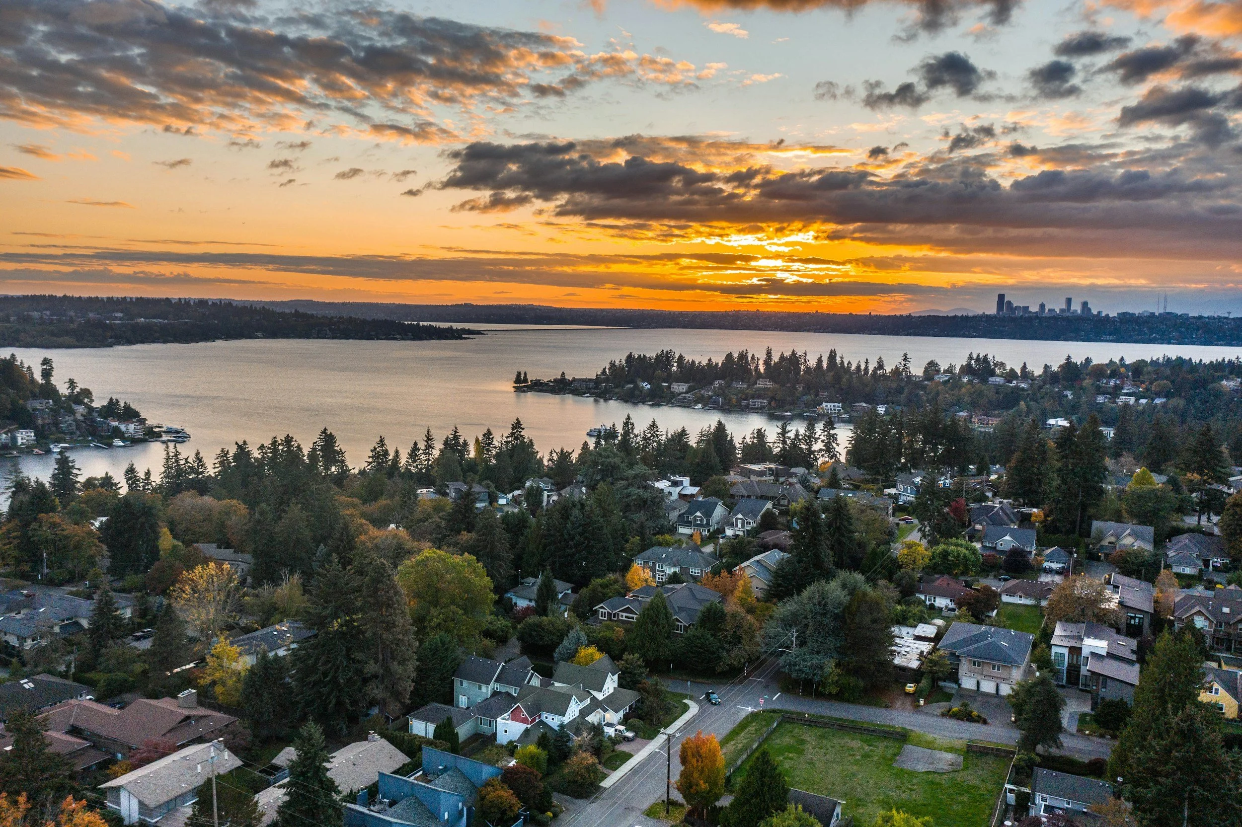An aerial view of a suburban neighborhood near a large body of water during sunset, with a city skyline in the distance. Houses are surrounded by trees with colorful autumn foliage.