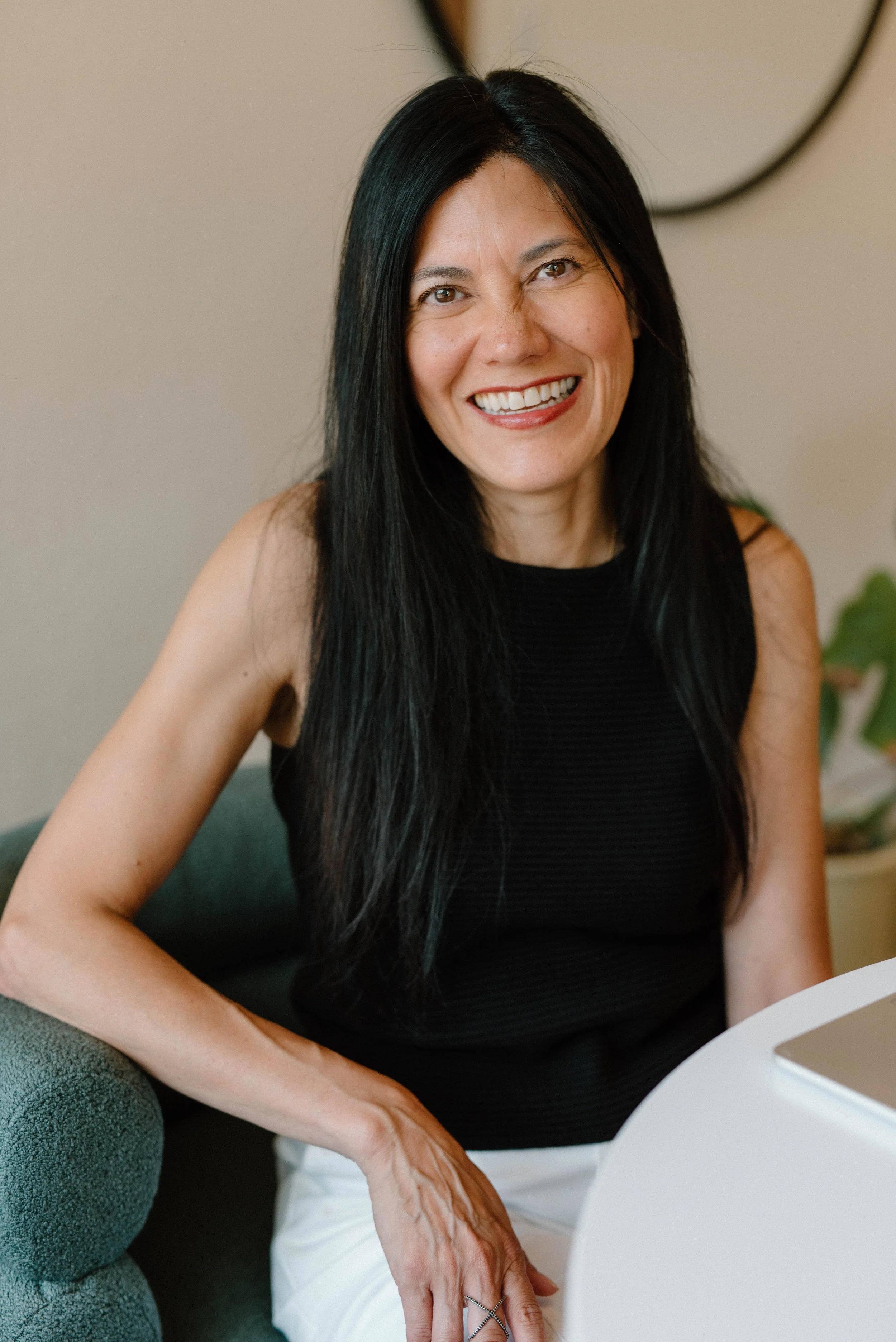 A woman with long black hair, smiling, seated indoors near a white table, wearing a sleeveless black top and white pants.