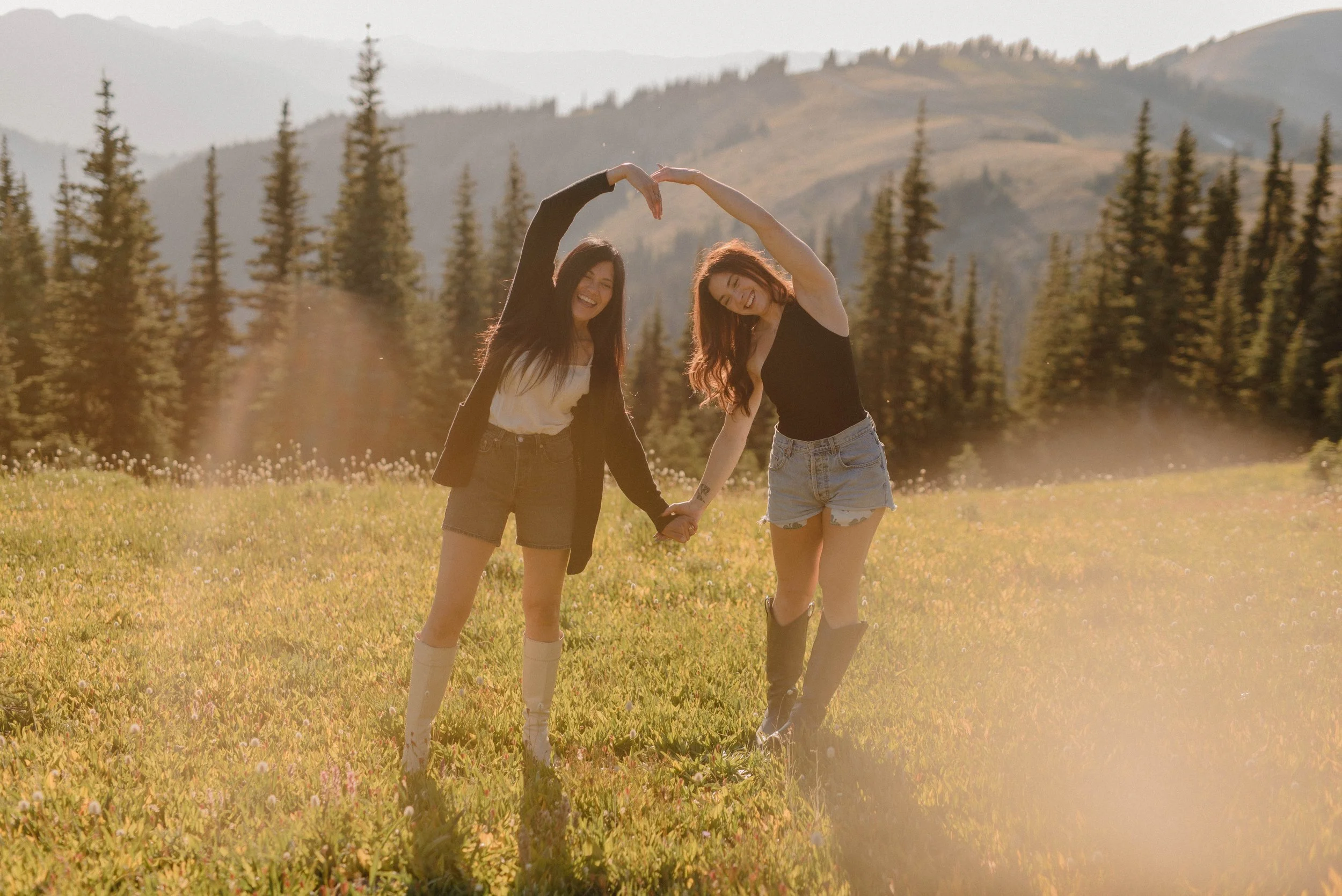 Two women holding hands and forming a heart shape with their arms in a grassy field with trees and mountains in the background during sunset.