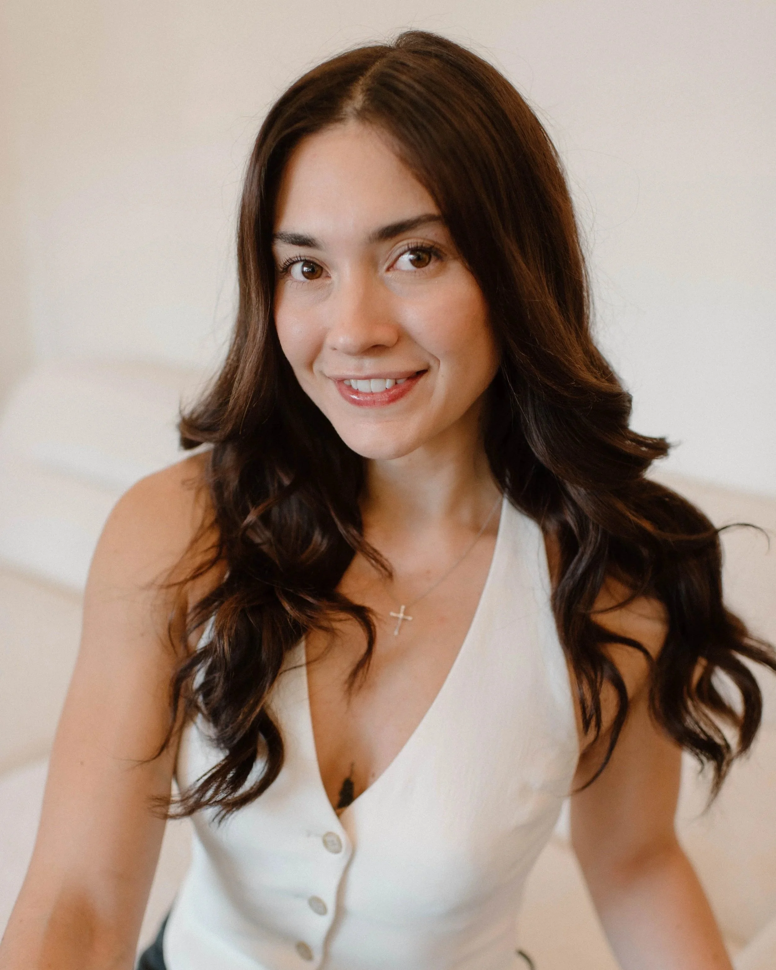 A young woman with long dark hair smiling at the camera, wearing a white sleeveless button-up top and a silver cross necklace, sitting on a white couch in a bright room.