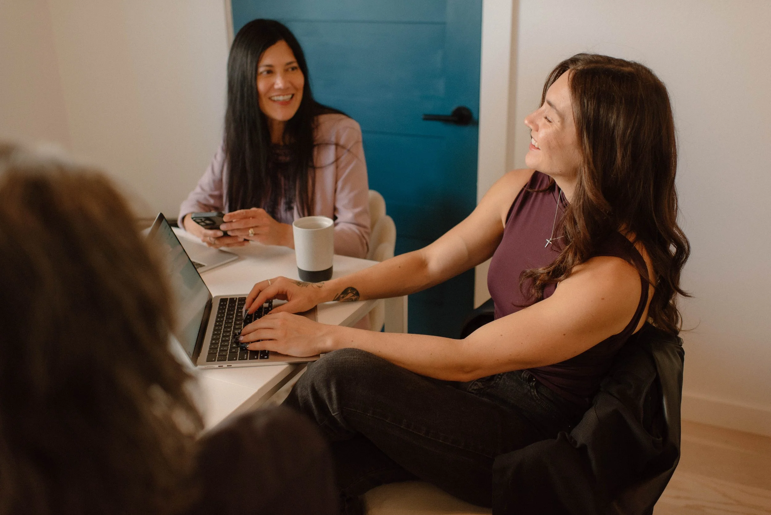 Three women sitting at a table smiling and talking; one woman is using a laptop, and another is holding a phone; a coffee mug is on the table.