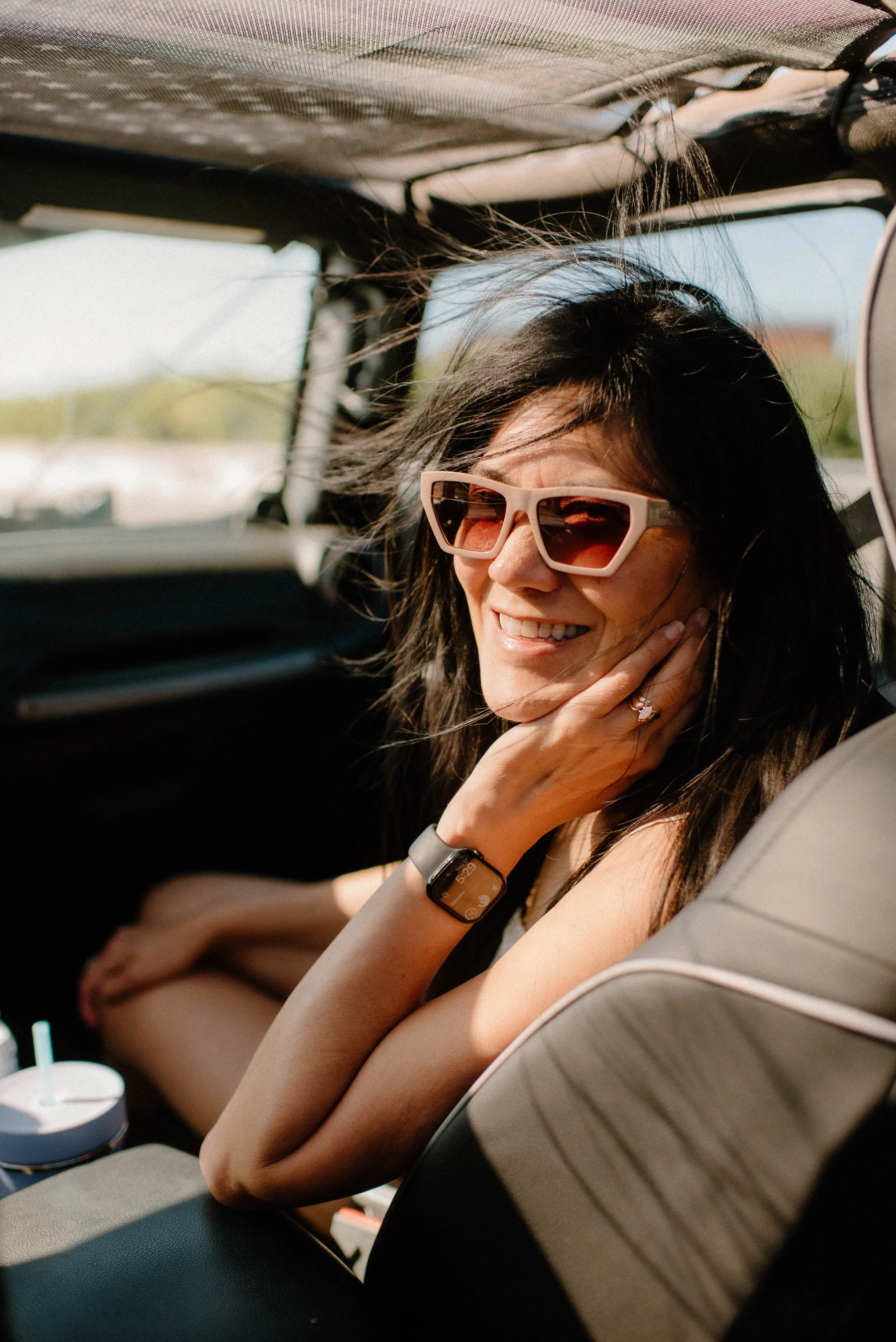 A woman smiling and wearing sunglasses sitting in the back seat of a car with her hand near her face, during daytime with sunlight.