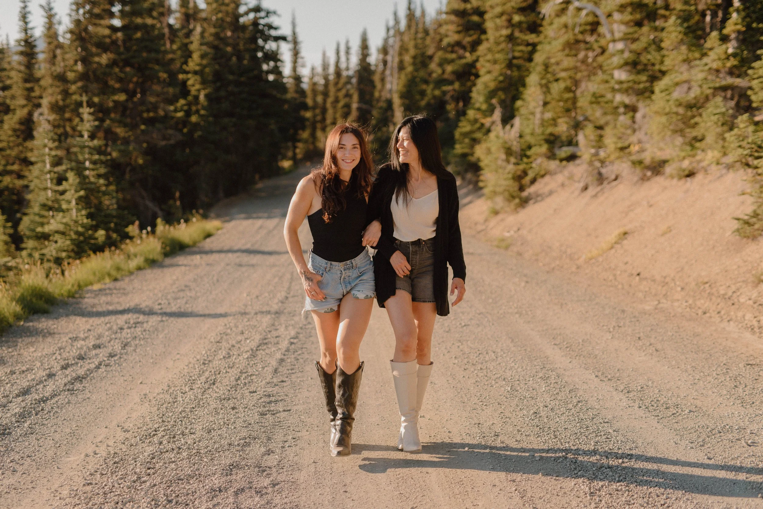 Two women walking together on a dirt road in a forested area during sunset, smiling and holding arms.