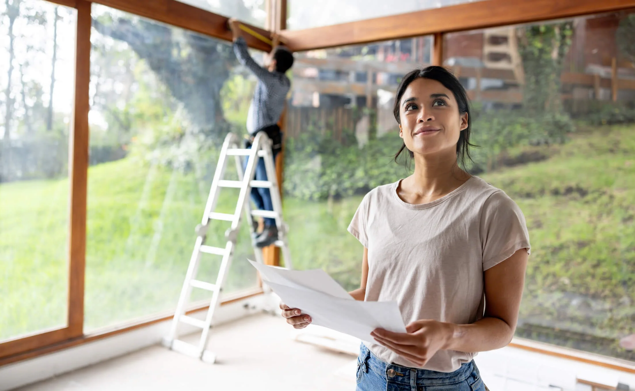 A woman holding papers inside a house with a large glass window behind her, while a man on a ladder works on installing or repairing the window.