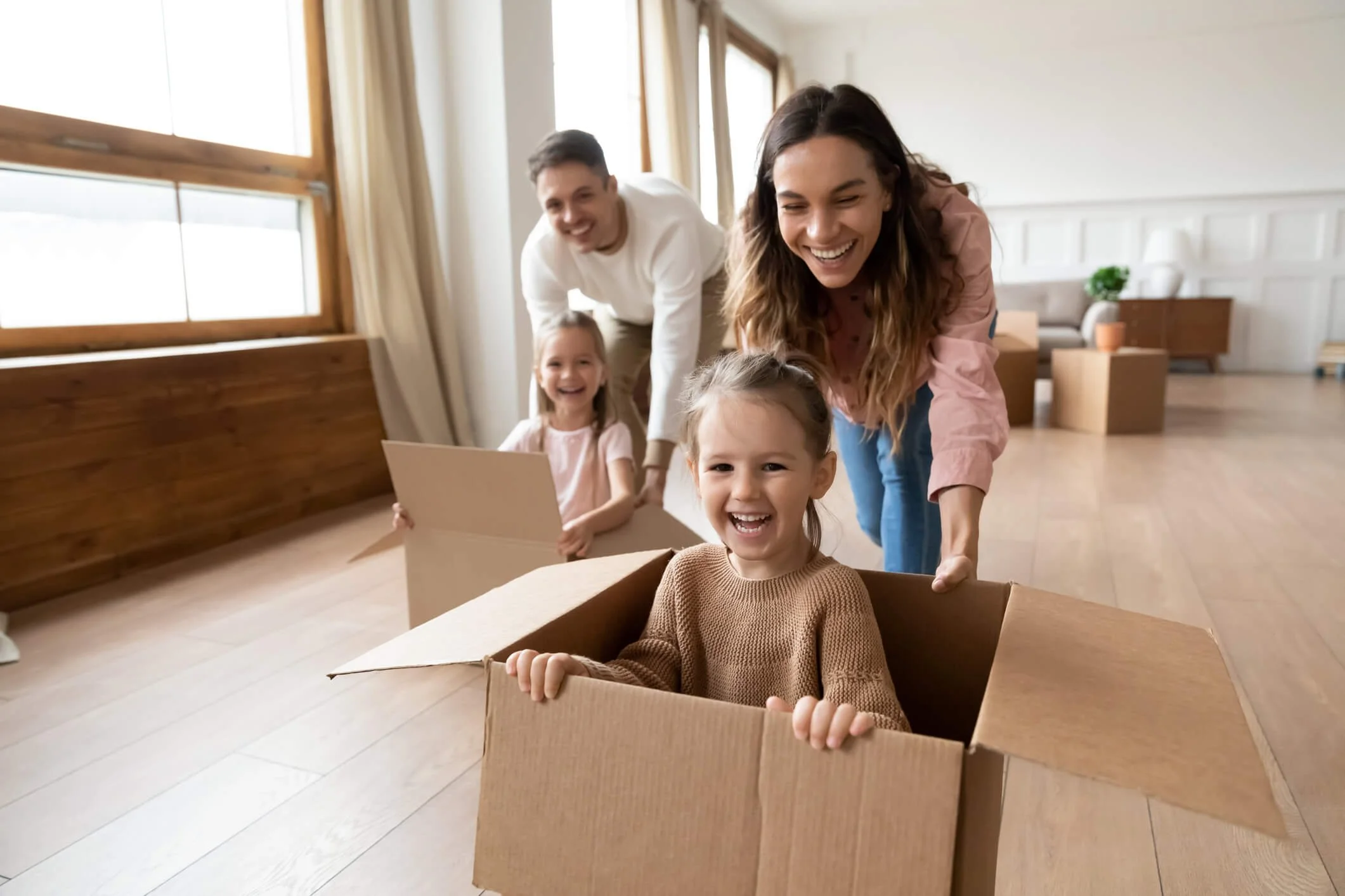 A happy family of four playing with a cardboard box inside a bright living room. The mother and father are pushing a little girl in a box, and another girl is standing behind them holding a piece of paper, all smiling and having fun.
