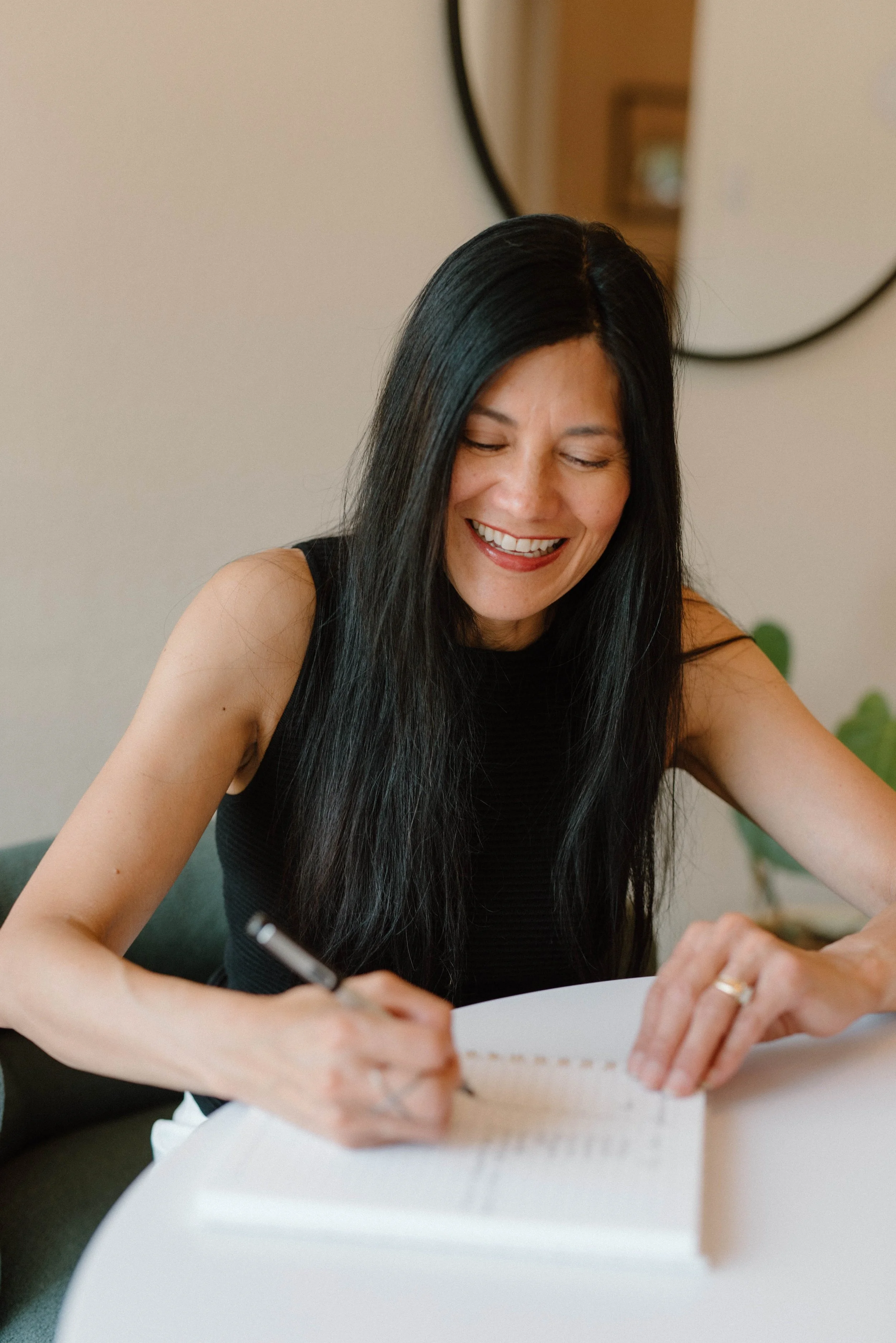 A woman with long black hair wearing a black sleeveless top, smiling while writing in a notebook at a white table.
