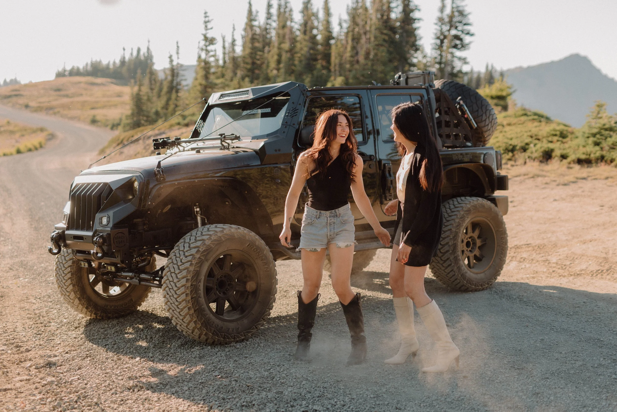 Two women standing beside a black off-road vehicle on a dirt trail in a mountainous area, smiling and talking to each other with trees and mountains in the background.
